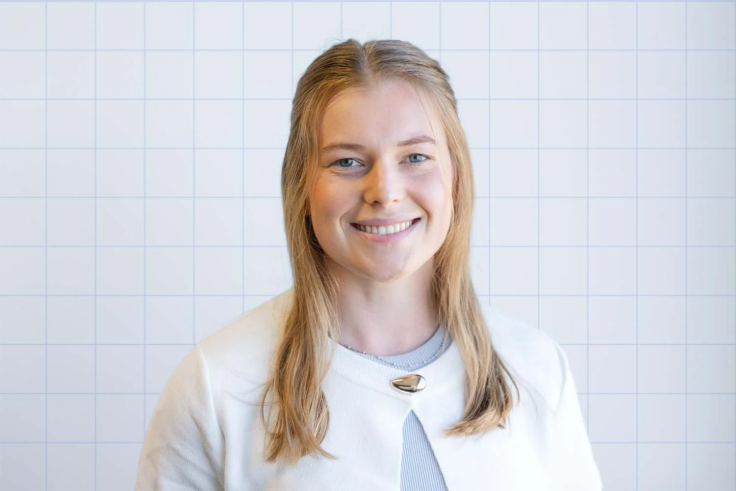 A woman with blonde hair and blue eyes smiling at the camera, standing in front of a white tiled wall.