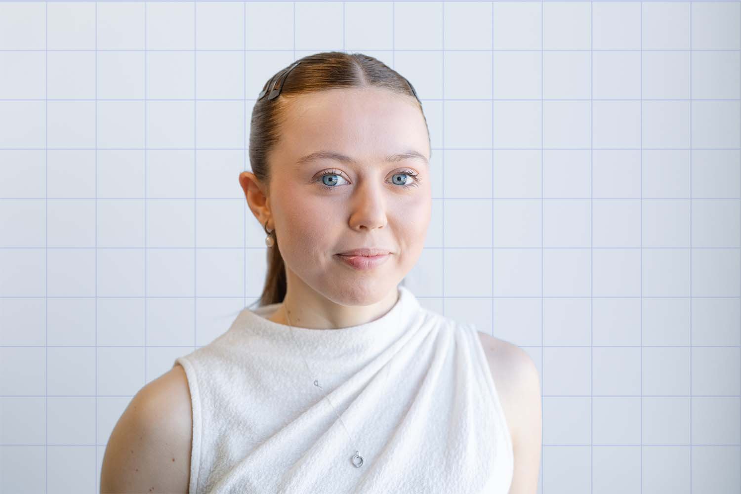 Grace at TIPS Financial Services in Perth portrait with blue eyes and light brown hair in a sleek ponytail, wearing a white sleeveless top and earrings, standing in front of a white tiled wall.