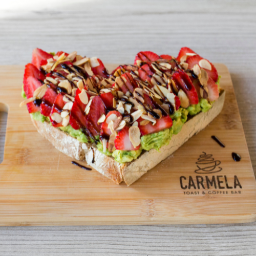 A heart-shaped toast topped with strawberries, sliced almonds, chocolate drizzle, and sliced pistachios, served on a wooden board with "Carmela Toast & Coffee Bar" lettering.