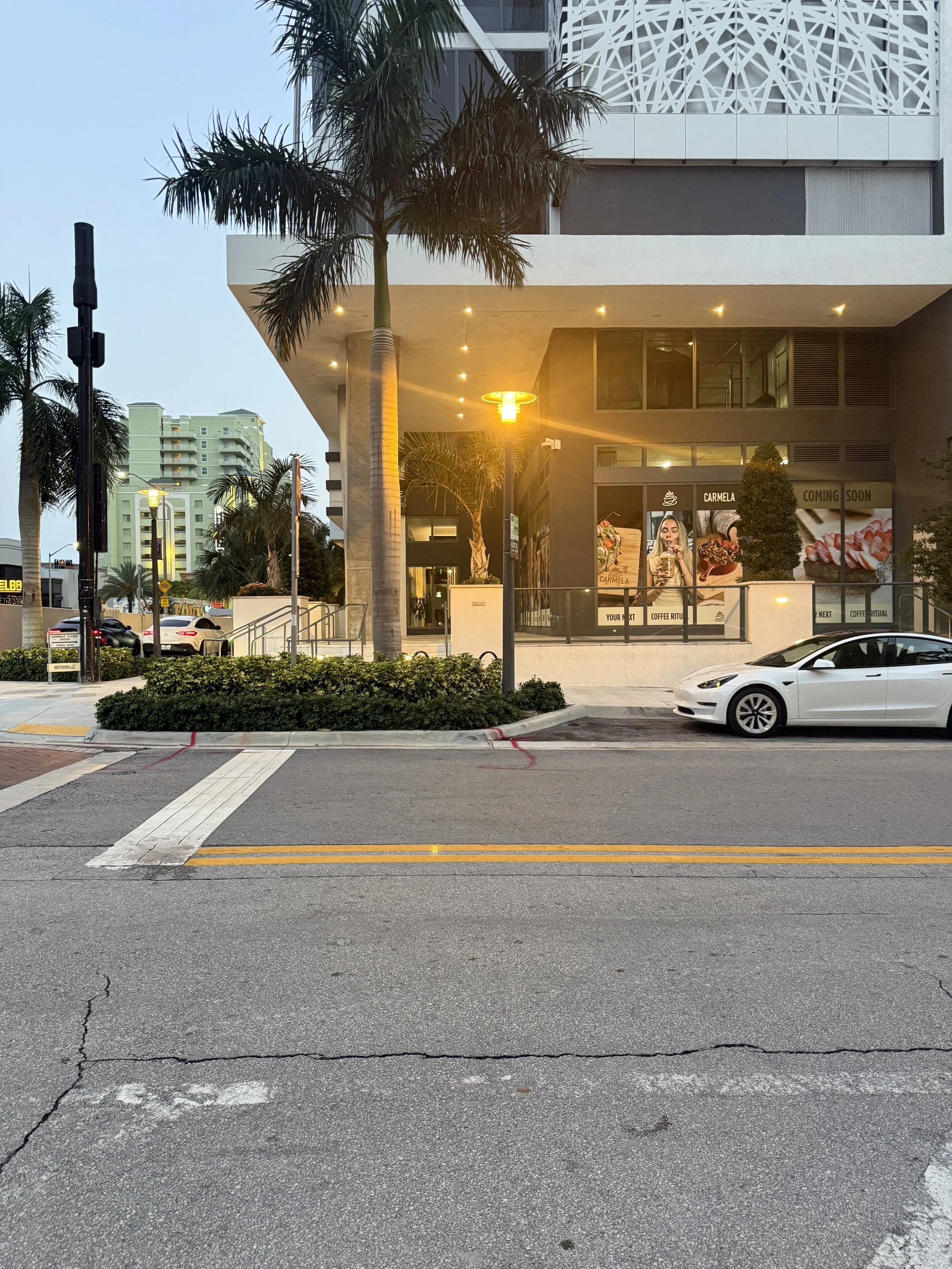 City street scene during dusk showing parked white car, palm trees, sidewalk, streetlights, and modern building with a decorative facade.