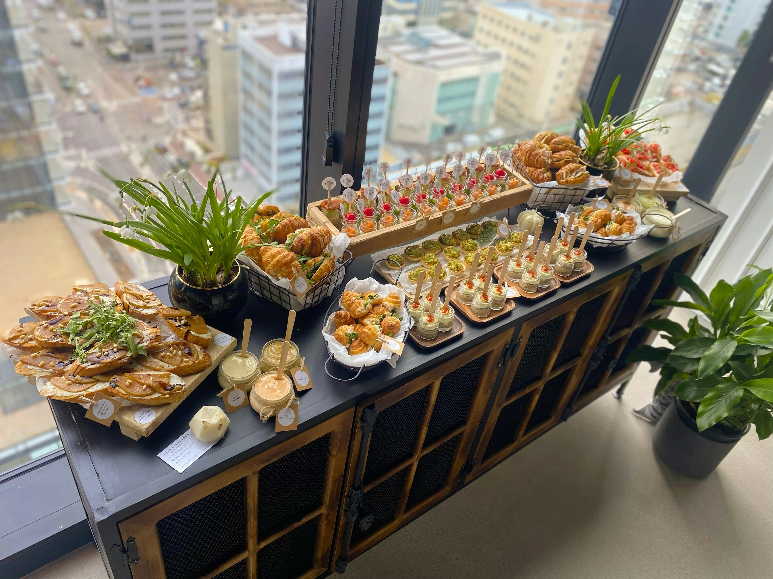 Table with an assortment of finger foods including pastries, small desserts with sticks, and plants, set in a high-rise building with a city view through the windows.