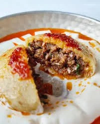 A close-up of a partially sliced empanada filled with seasoned ground meat, served on a white plate with red and orange sauce drizzles.