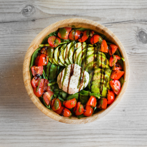 A wooden bowl containing a fresh salad with sliced avocado, cherry tomatoes, sliced cucumbers, and a ball of burrata cheese drizzled with balsamic glaze, placed on a light wooden surface.
