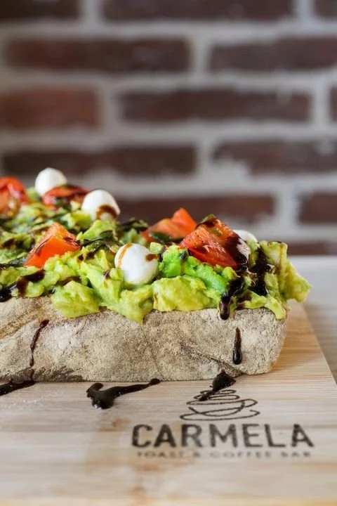 A slice of avocado toast on a wooden board with tomatoes, mozzarella, and drizzled with balsamic glaze. The background features a brick wall.