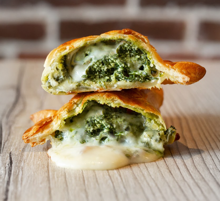 Close-up of a sliced savory pastry filled with melted cheese and spinach, placed on a wooden surface with a brick wall background.
