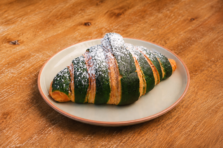 A croissant with green icing, topped with powdered sugar, on a white plate on a wooden table.