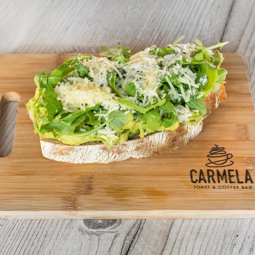 Open-faced avocado toast topped with arugula and shredded cheese on a wooden cutting board that says Carmela Toast & Coffee Bar.