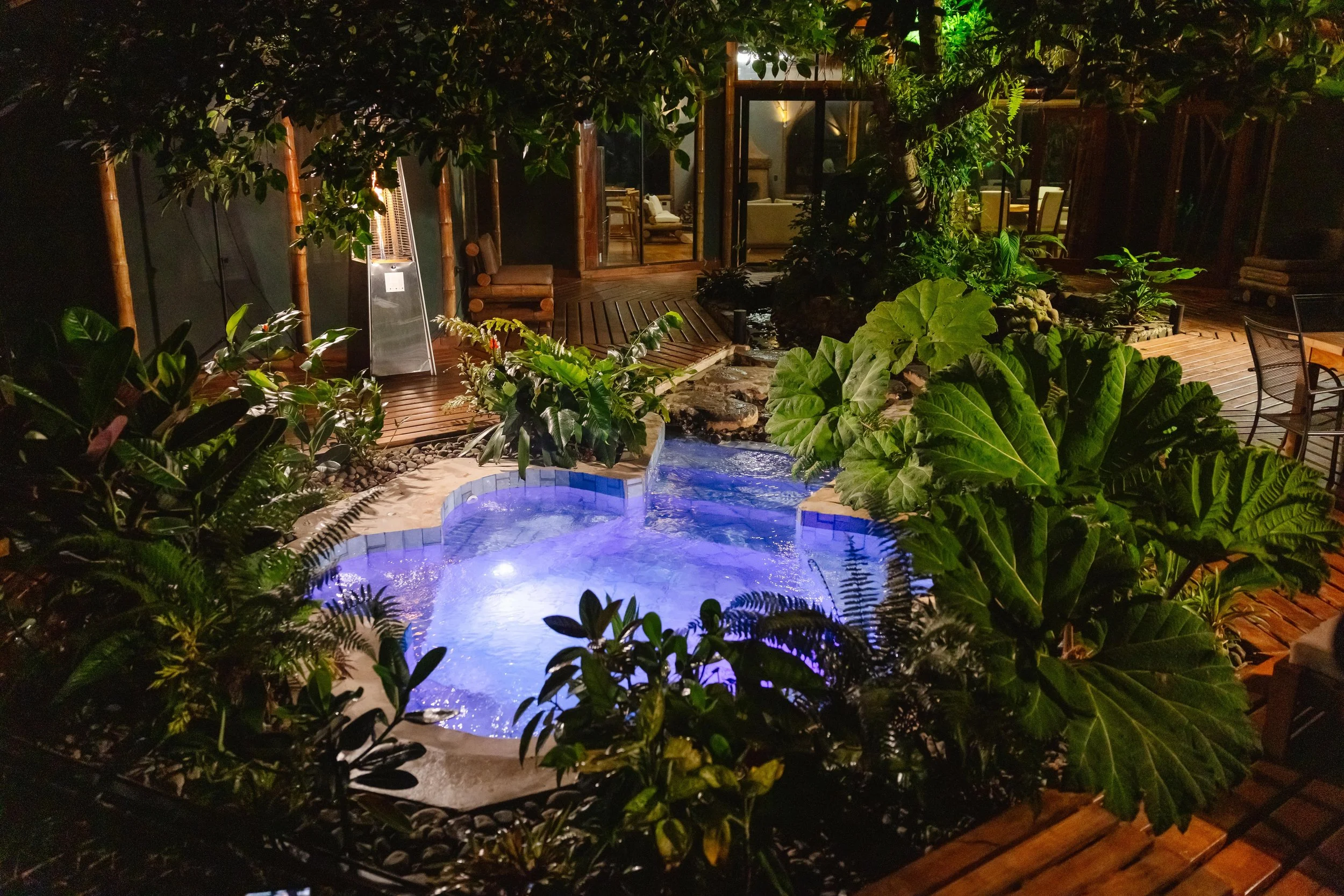 Nighttime view of an outdoor patio with lush green plants, a small illuminated hot tub, and a wooden deck leading to a glass door.