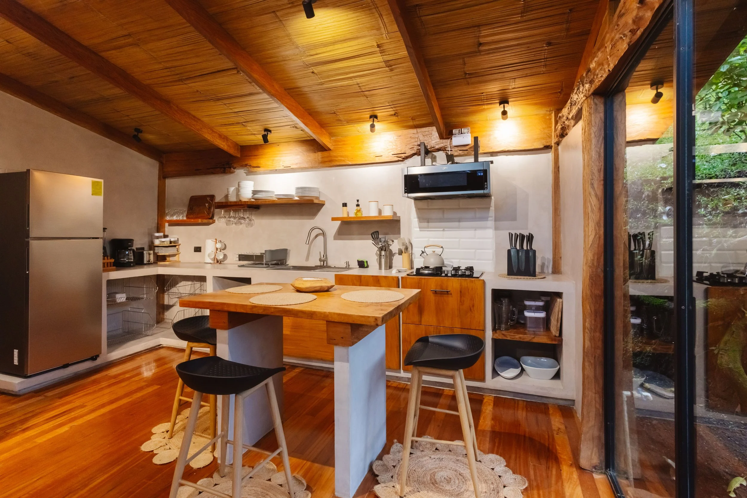 Warm wooden kitchen at El Tigre Hideaway with open shelving, stainless-steel refrigerator, coffee maker and a large window facing lush Monteverde greenery.