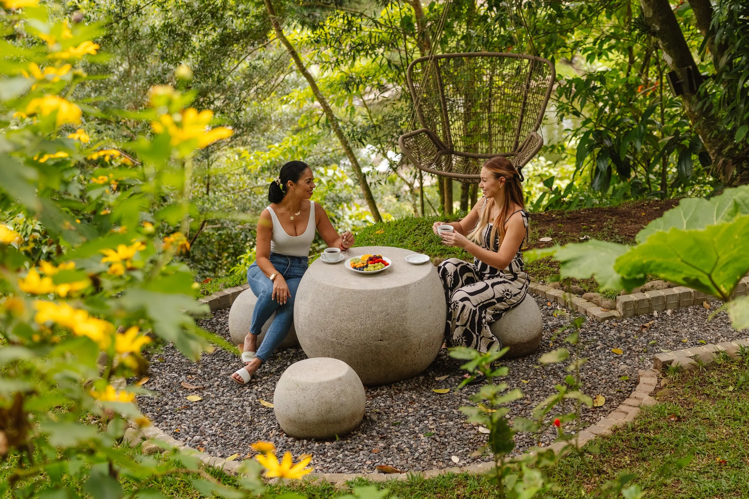 Two women sitting at a round outdoor table in a lush garden, having coffee and conversation. A hanging chair is in the background, surrounded by green foliage and yellow flowers.