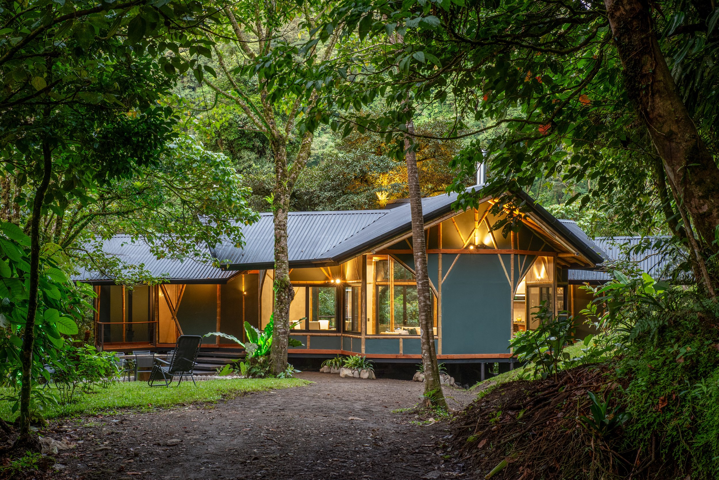 Modern house with large glass windows and a metal roof, set in a lush, green forest with trees and plants surrounding it. Outdoor furniture is visible in the yard, and the house is illuminated from inside, giving a cozy appearance.