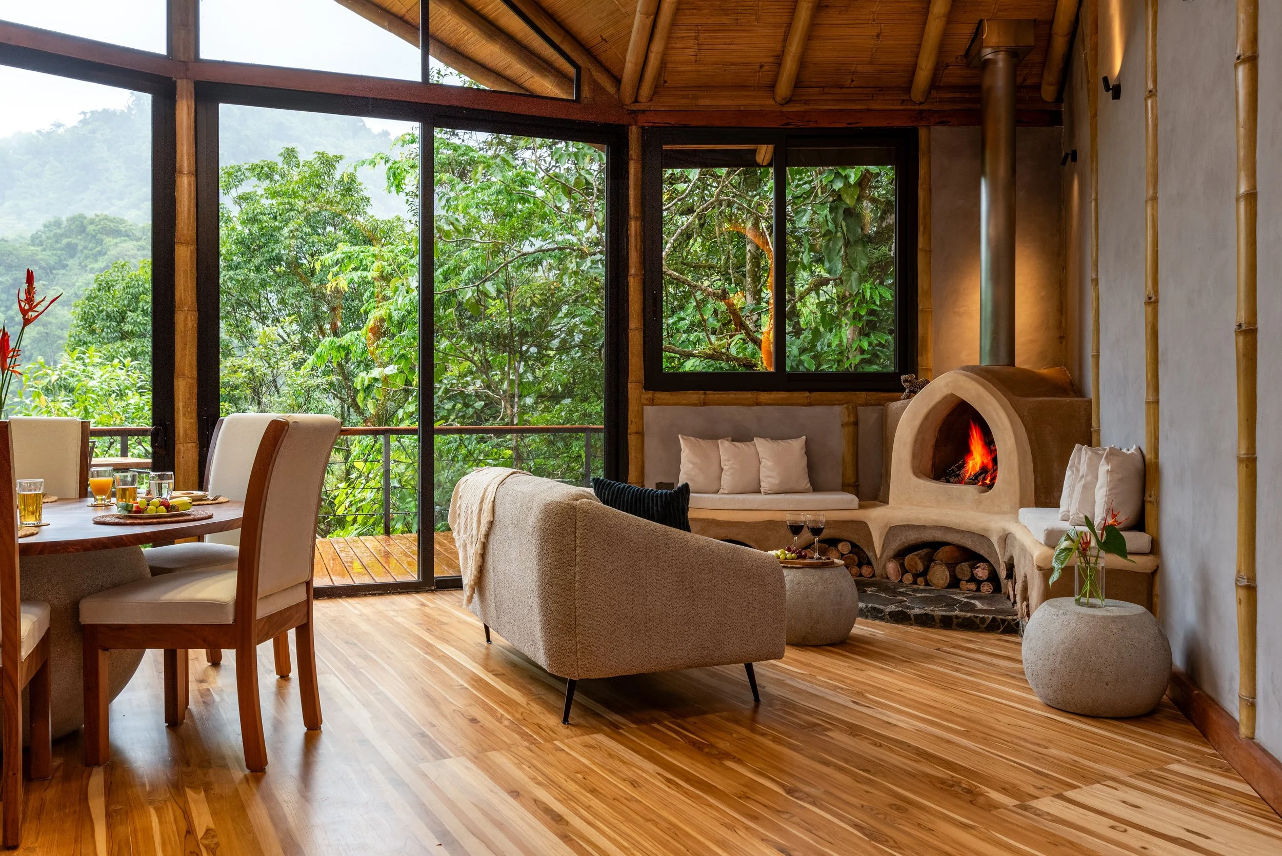 Living room with large windows showing lush green trees and mountains outside, wooden ceiling, and bamboo accents. There is a beige sofa, a cozy fireplace with logs, and a dining table set for breakfast with glasses, juice, and fruit.