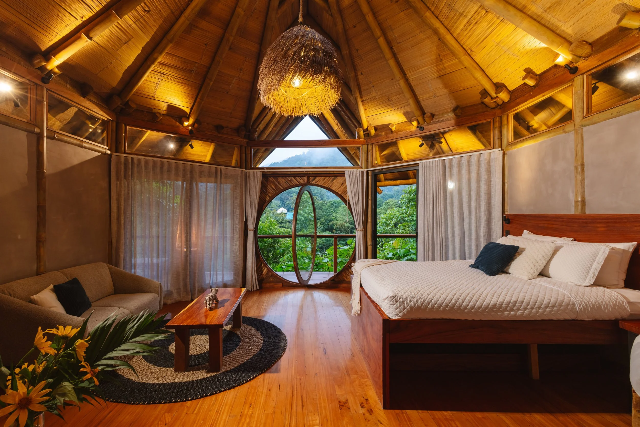 Bedroom with bamboo walls and ceiling, large window with mountain view, king-size bed, beige couch, wooden coffee table, floral arrangement, warm lighting, and rustic decor.