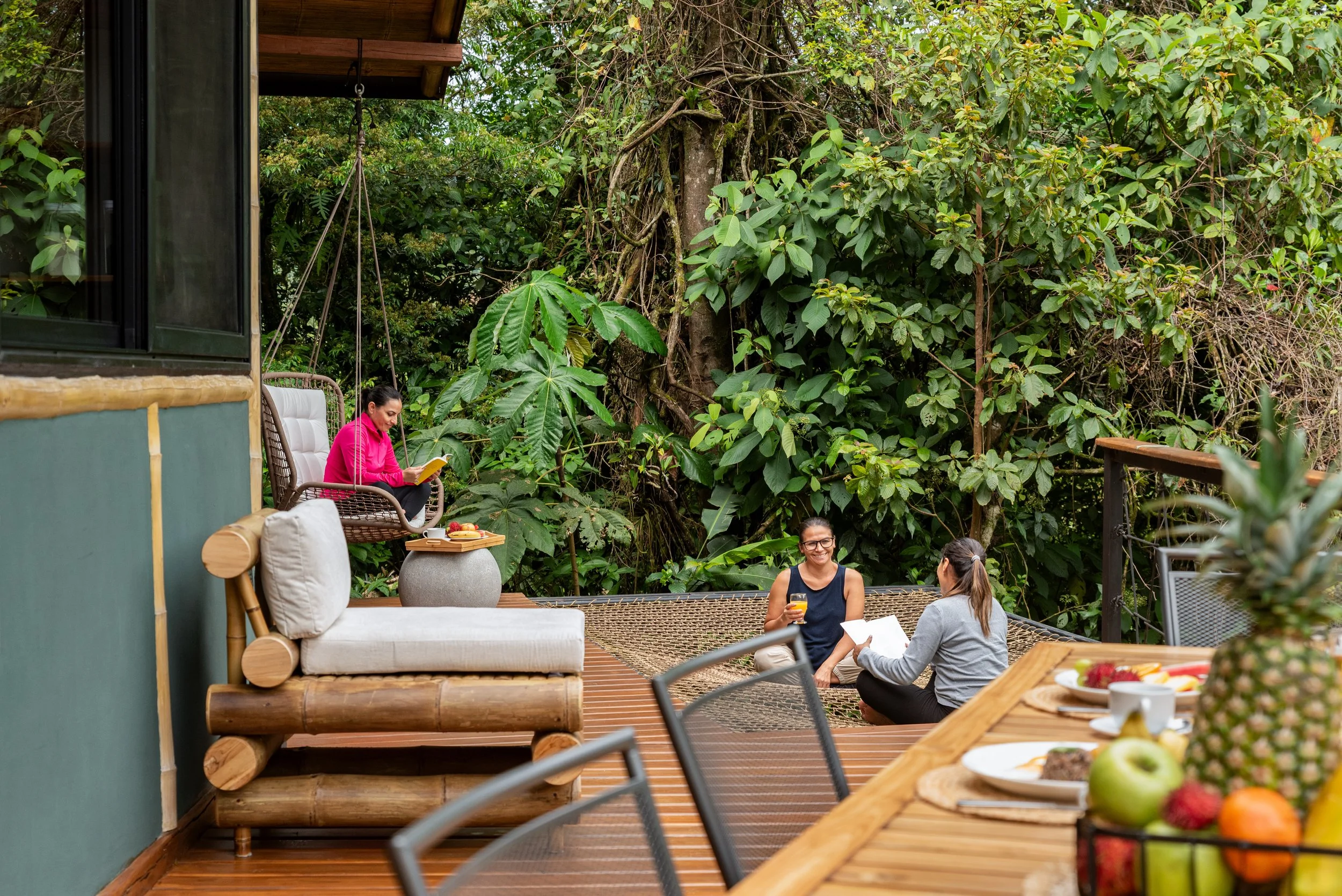 People enjoying a gathering outdoors on a wooden deck surrounded by lush green foliage. Some are sitting on chairs, chatting and drinking, with a table of food in front, including fruit and pastries. One woman is reading, and others are smiling and t