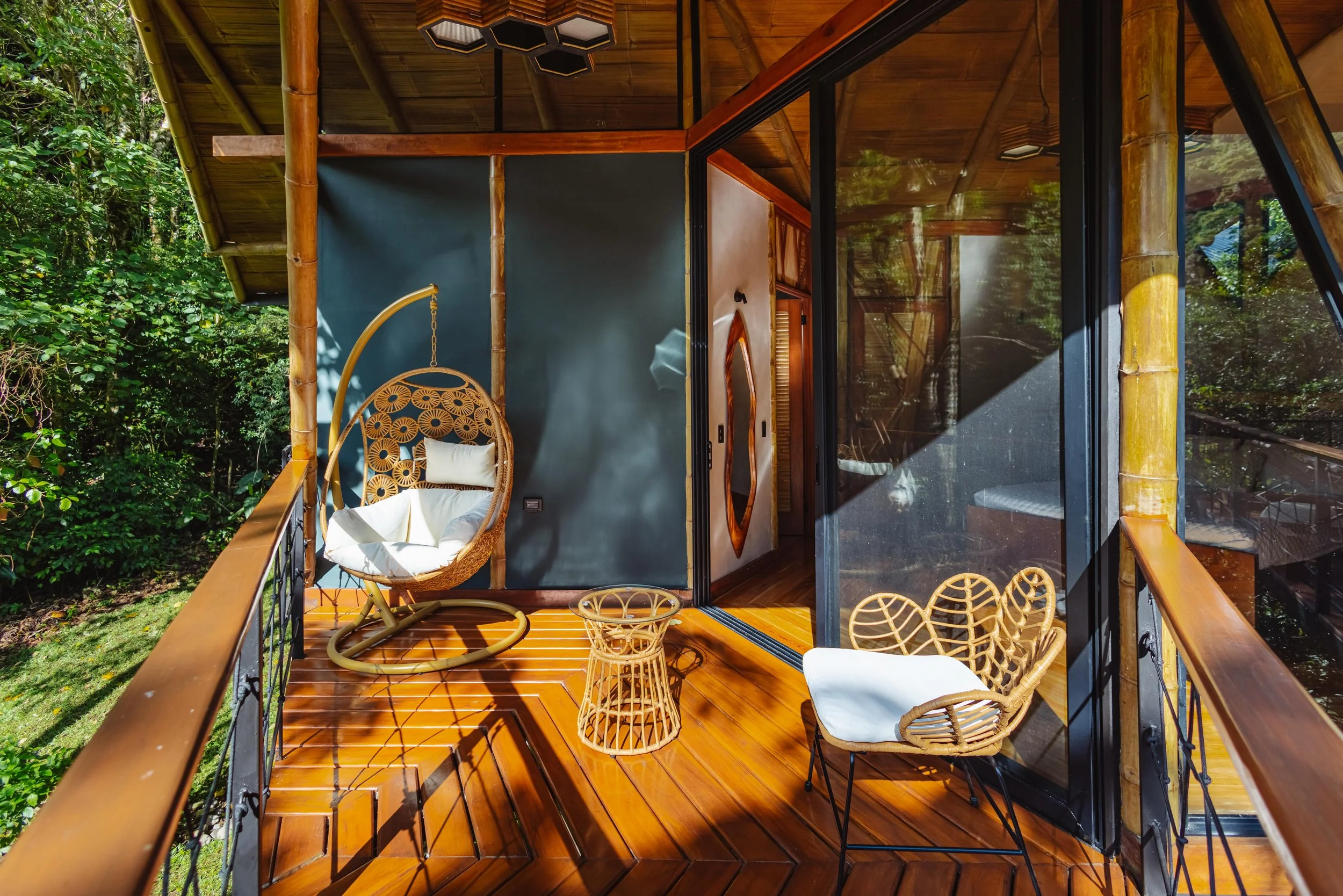 Balcony of a wooden cabin with wicker chairs, a hanging swing chair, and glass sliding doors reflecting trees and sky, surrounded by lush greenery.