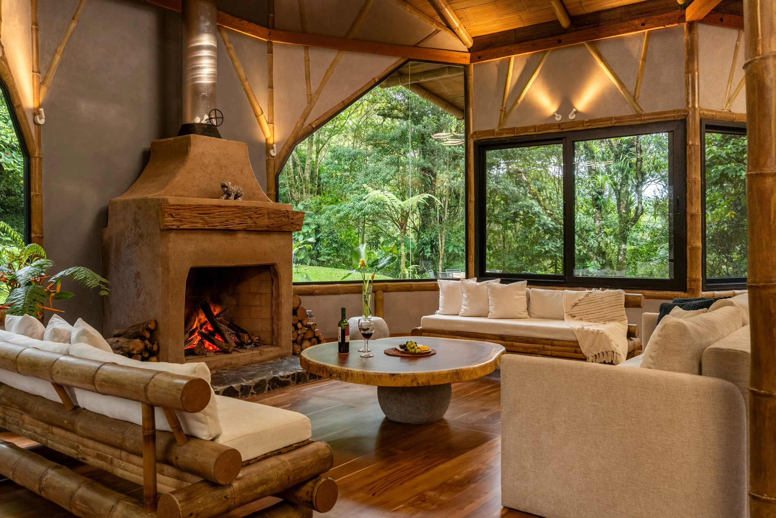 A cozy living room with bamboo furniture, a fireplace with a fire burning, large windows showing lush green trees outside, and a round wooden coffee table with a bottle of wine, a glass of wine, and a plate of fruit.