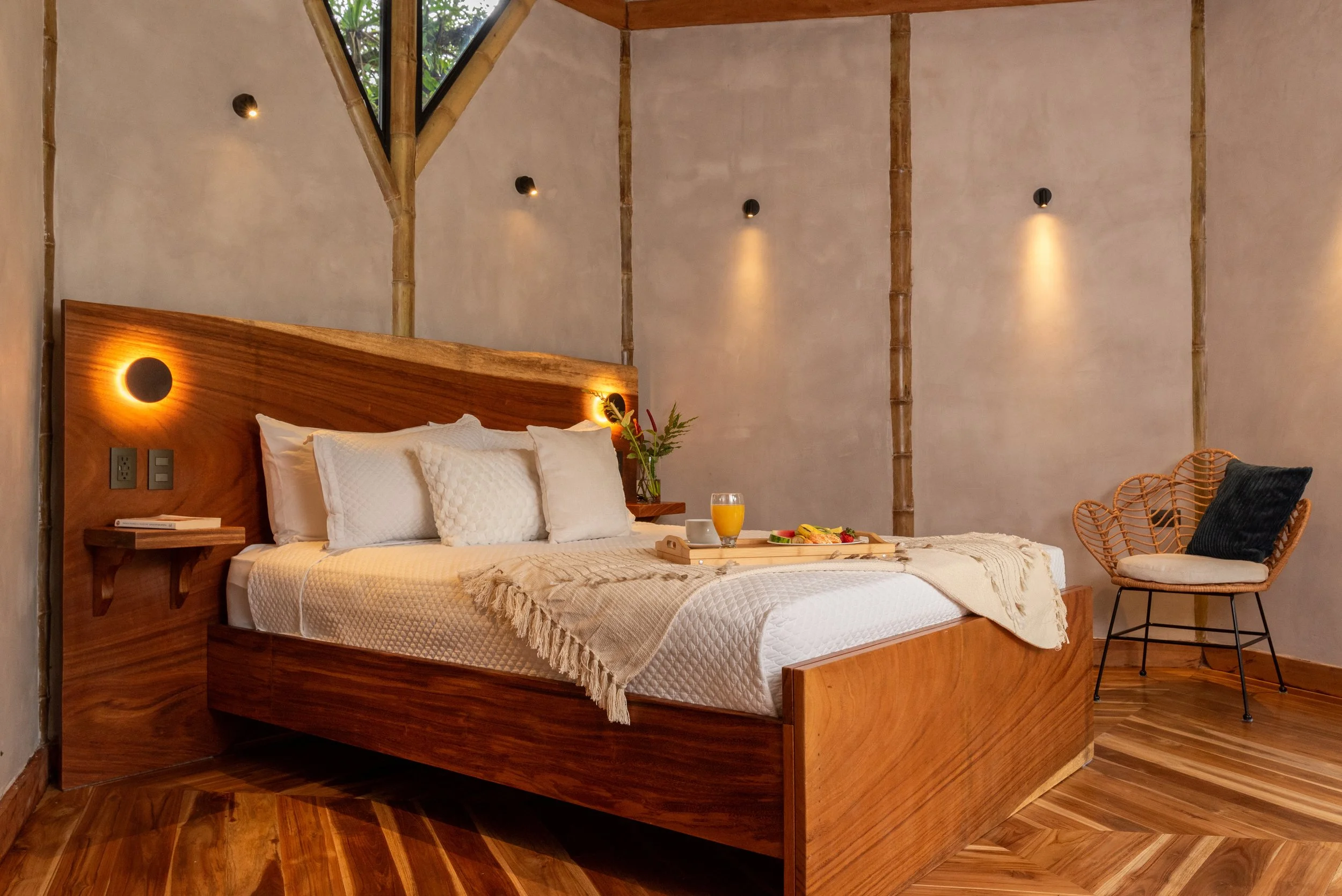 A cozy bedroom with a wooden bed, white bedding, and decorative pillows. A bedside table with a book and a glass of orange juice. Wicker chair with cushion, plants, and modern wall-mounted lights create a warm, inviting atmosphere.