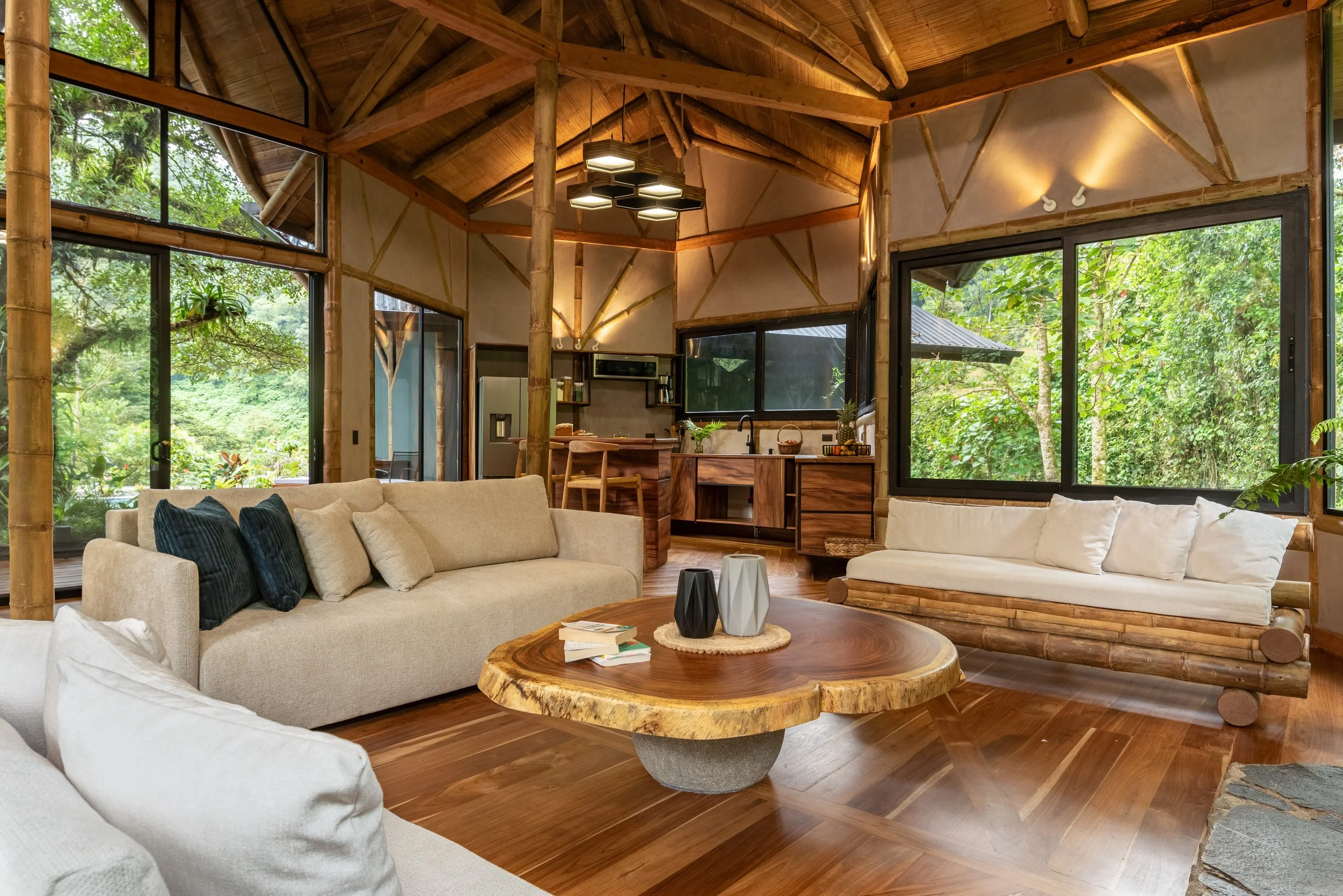 Living room with wooden ceiling beams, large windows showing green trees outside, beige sofas with dark and white pillows, a wooden coffee table with three black and white vases, and a kitchen area with a refrigerator, shelves, and a sink in the back