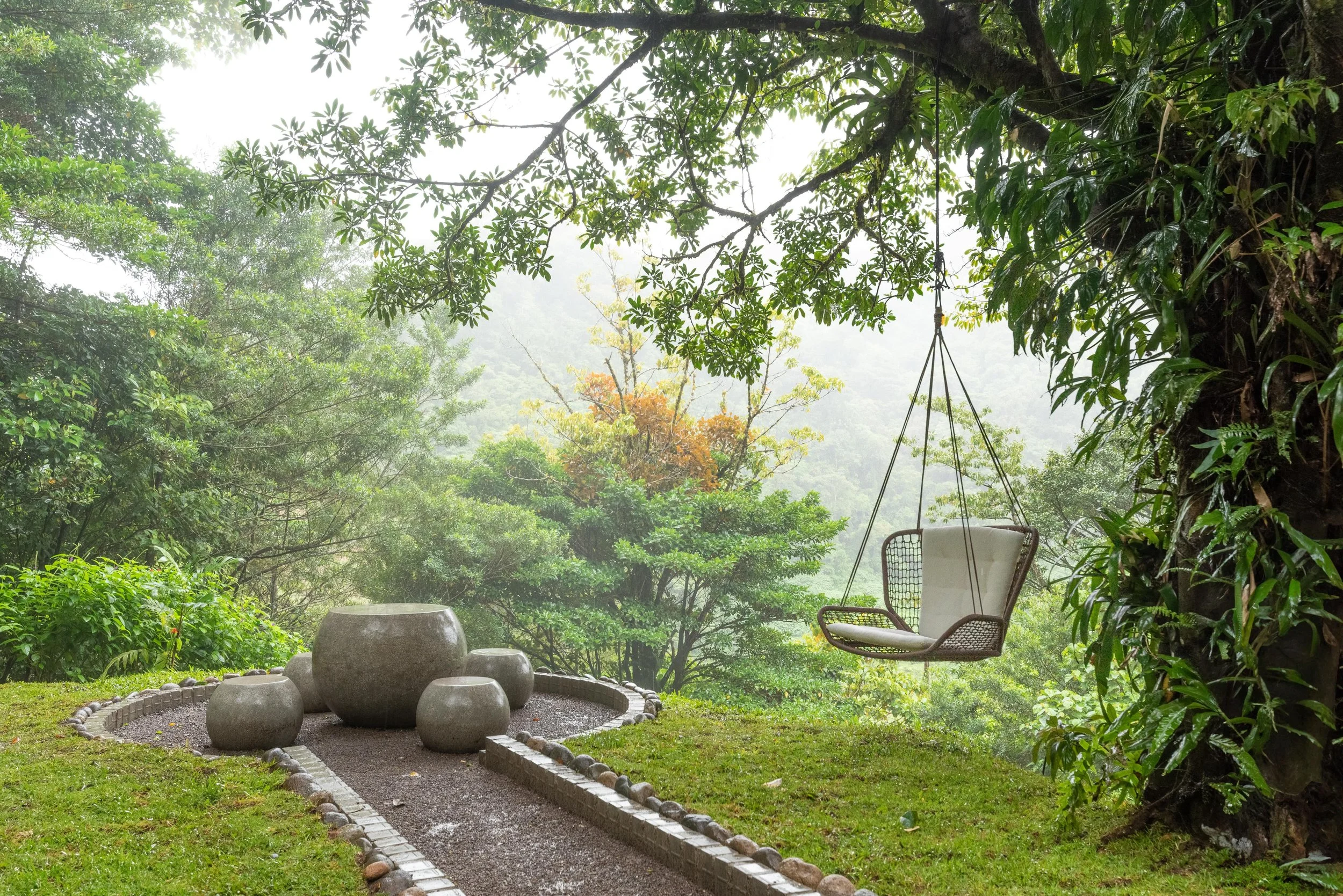 A garden scene with lush green trees and bushes, a gravel pathway bordered with bricks, a swing hanging from a tree, and large decorative stone pots arranged on the ground.