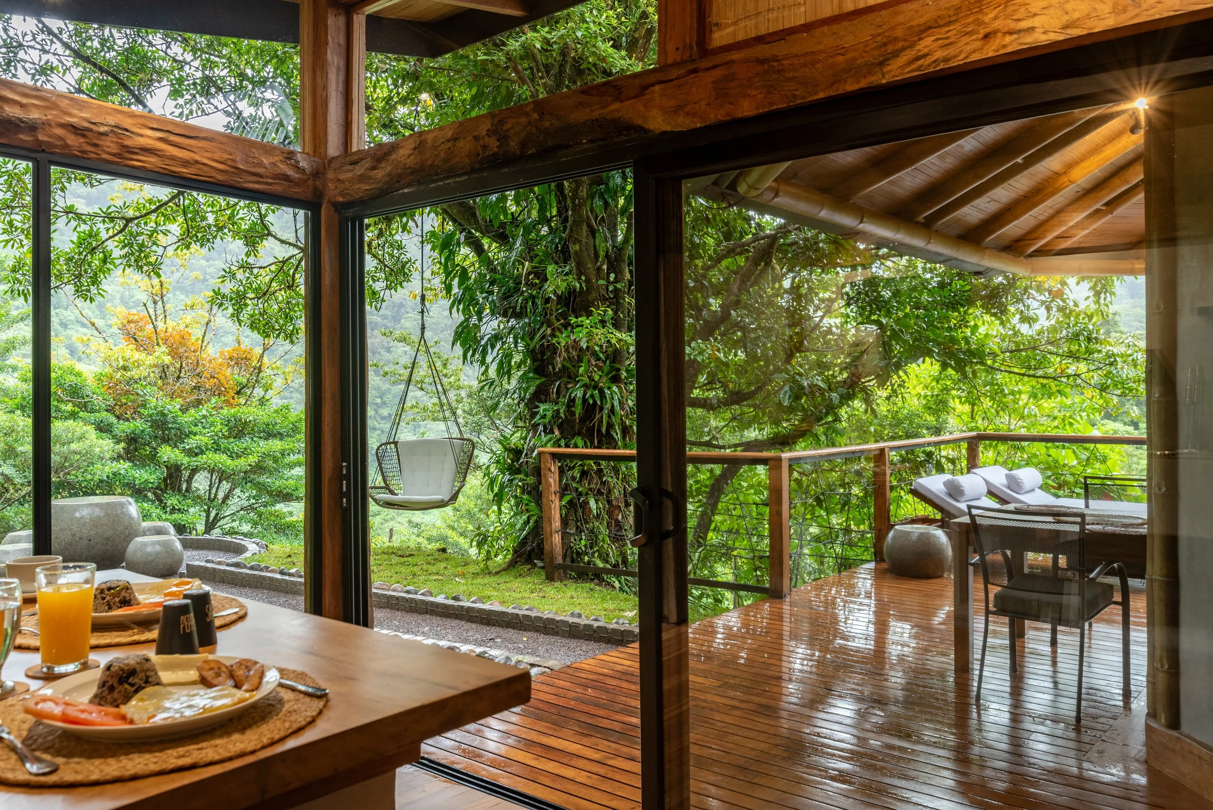 A dining area with a view of a green, wooded outdoor space through large glass doors. Part of a breakfast spread including eggs, baked goods, and drinks is visible in the foreground.