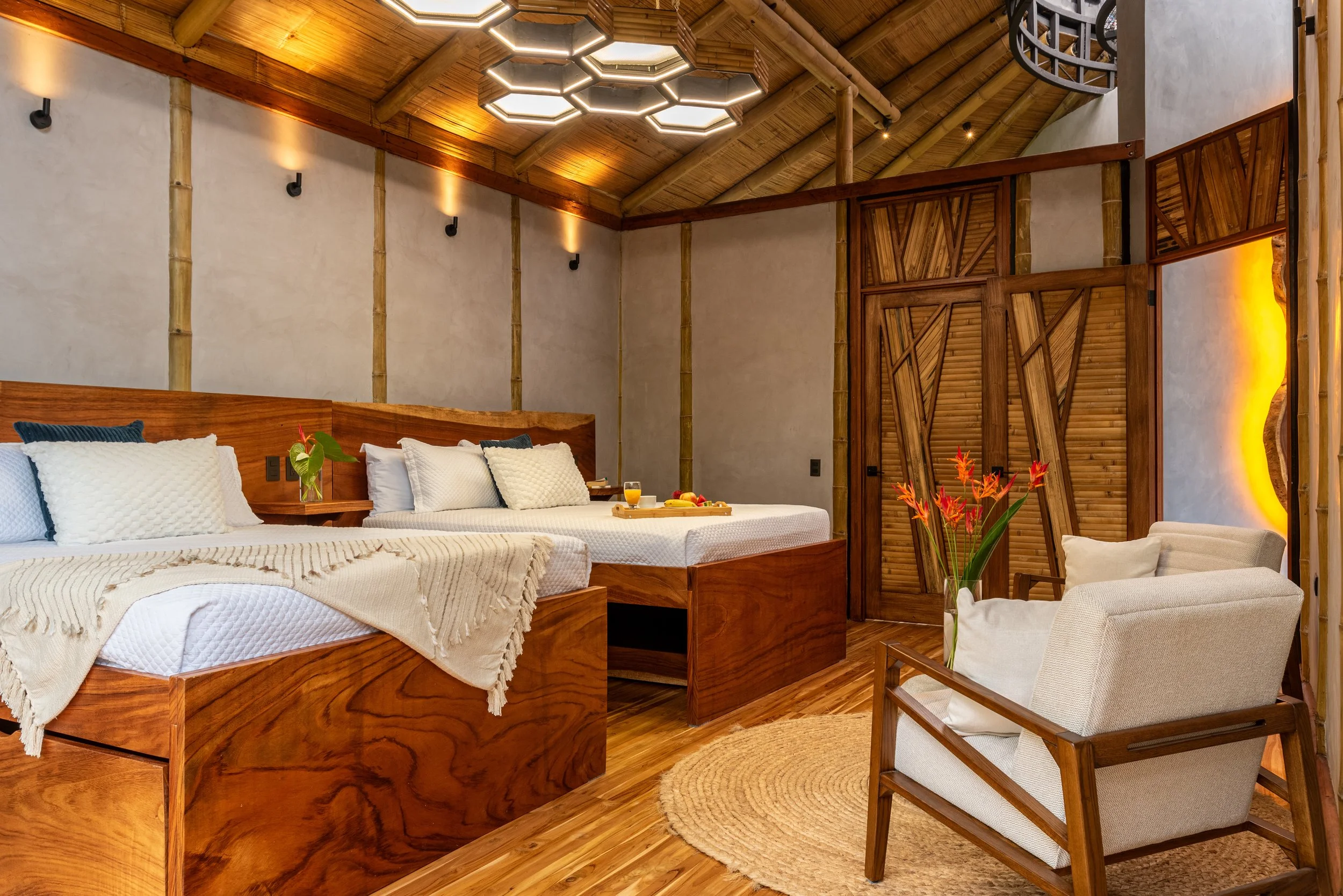 Interior of a bedroom with a high thatched wooden ceiling, wooden doors, and warm lighting. The room features a large wooden bed with white bedding, a wooden tray with food and drinks, a beige armchair, and tropical decor.
