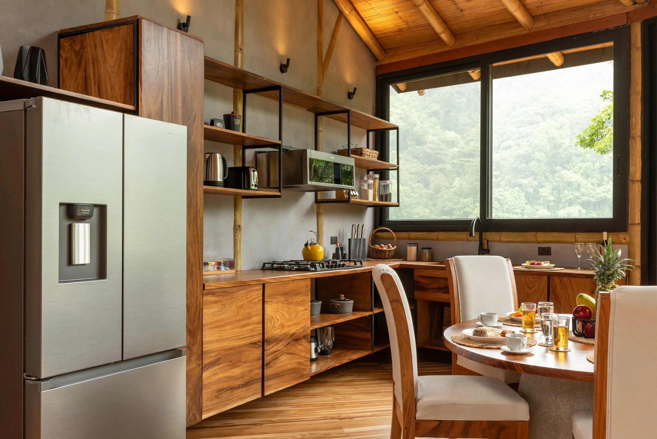 A modern kitchen with wooden cabinets, open shelving, a stainless steel refrigerator, and large windows offering a view of green trees outside. The kitchen has a cozy, natural aesthetic with contemporary appliances.