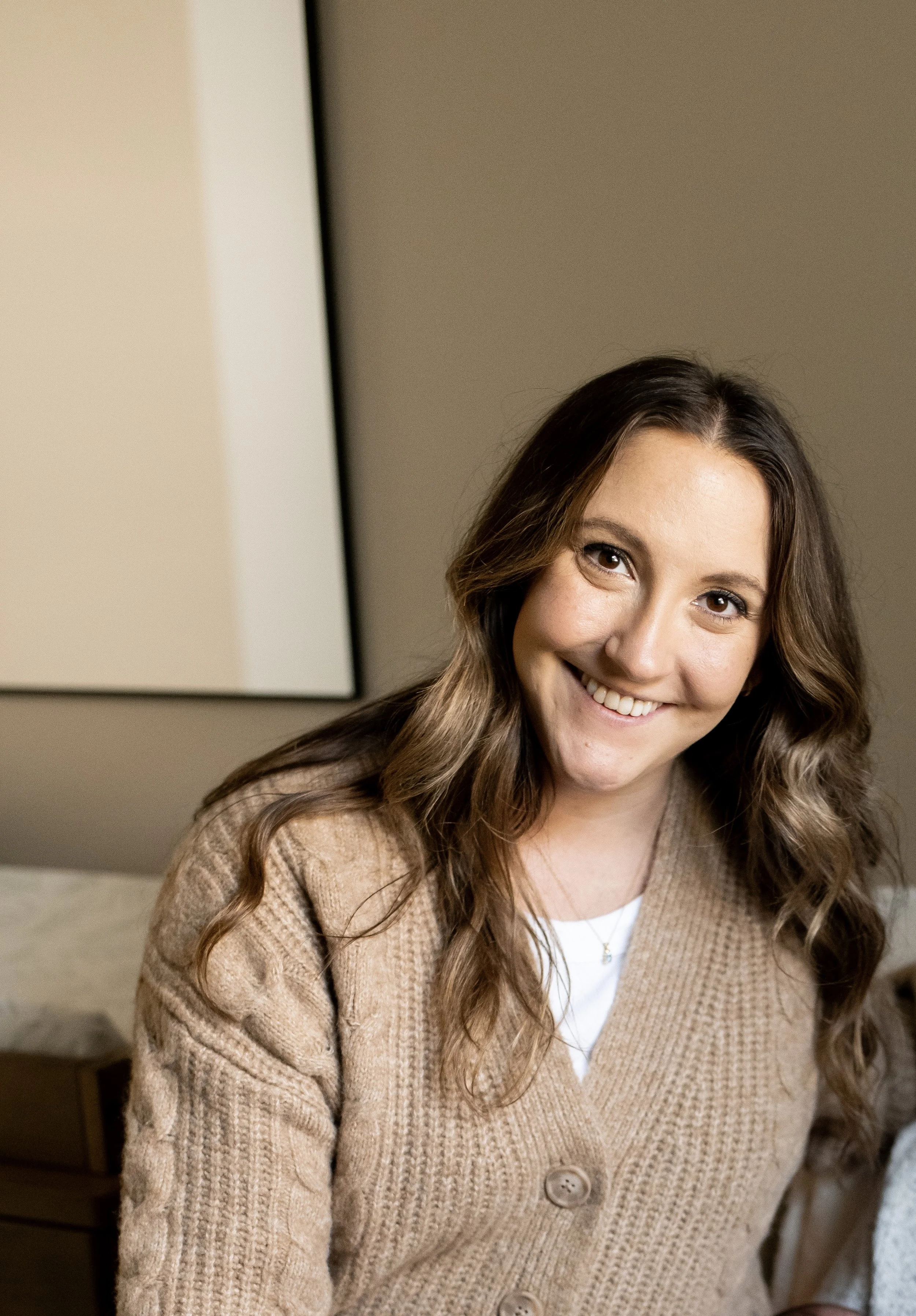 A woman with long wavy brown hair, smiling, wearing a beige cable-knit cardigan with large buttons and a white shirt underneath, sitting indoors.