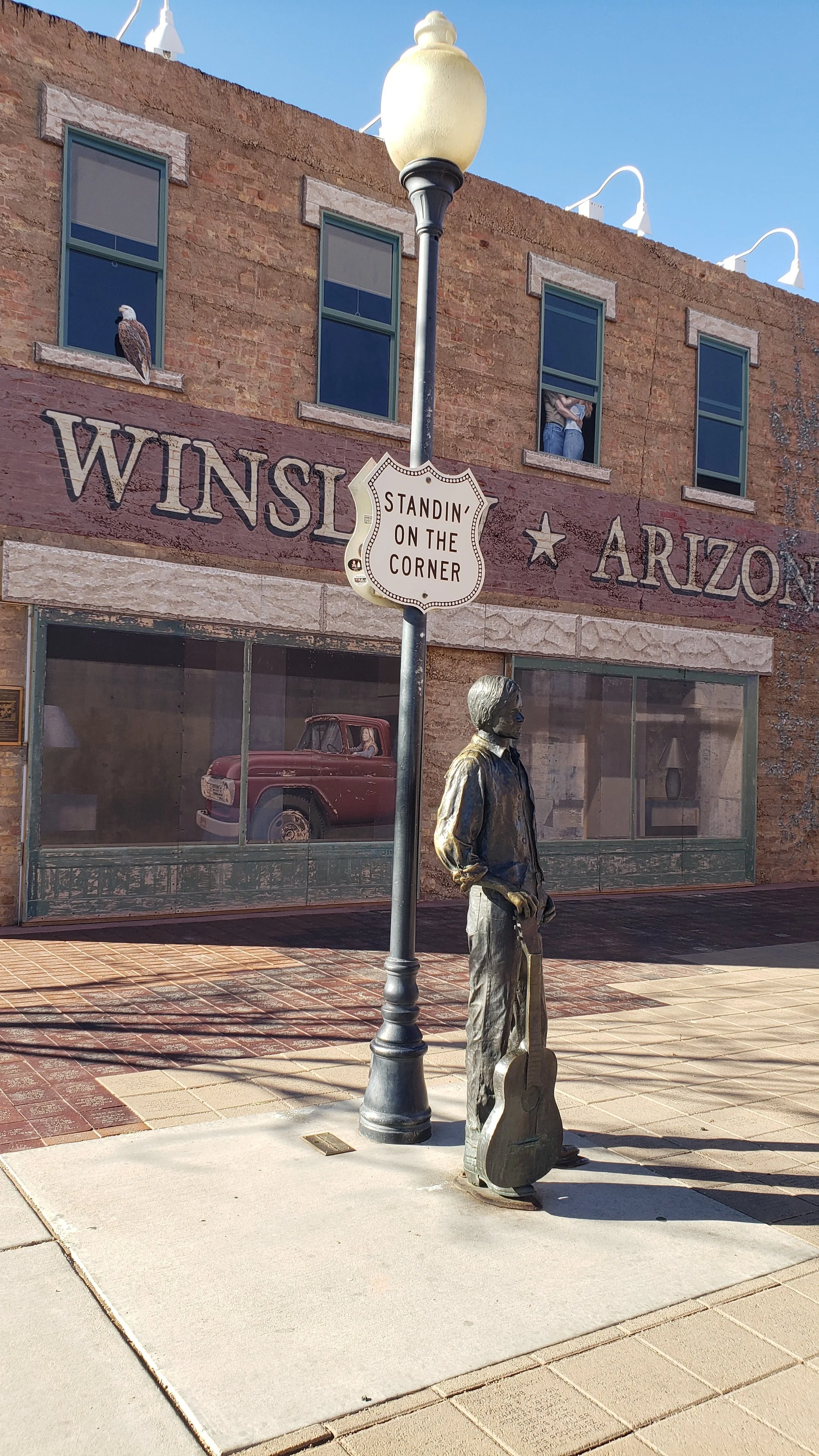 A street scene with a bronze statue of a young man holding a guitar, standing on a sidewalk near a lamp post. The lamp post has a sign that says "Standin' on the Corner." Behind, a brick building features a mural with a vintage car and three windows, one with a person looking out and others with people partially visible inside. An eagle perches on the window ledge of the building. The sky is clear and blue.