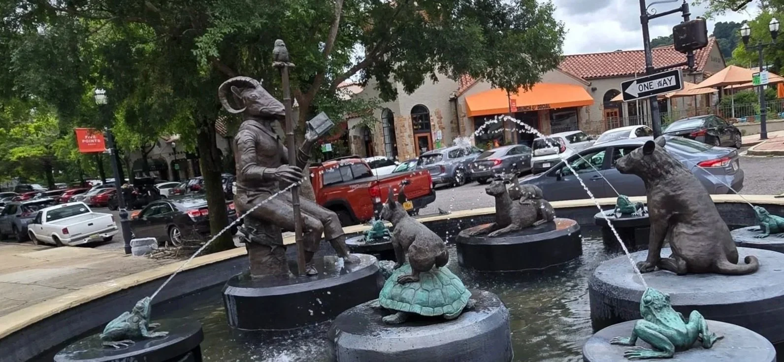 Public fountain featuring sculptures of animals and a person reading a book, with water spouting from the animals' mouths and hand in the background, cars parked along the street, and buildings with trees and street signs.