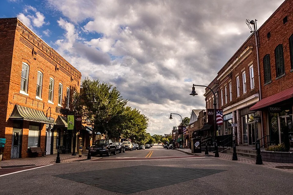 Main street view with brick buildings, parked cars, street lamps, trees, and cloudy sky in a small town.