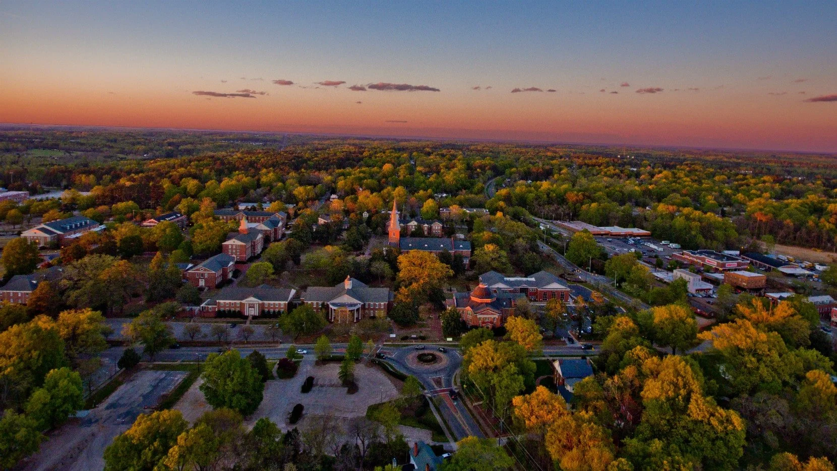 An aerial view of a college town during sunset, showing campus buildings, a church with a tall steeple, surrounding trees with fall colors, and commercial area with parking lots.