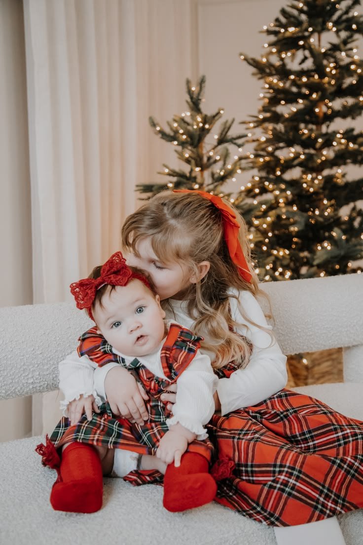 Two young girls in Christmas outfits sitting on a white bench next to decorated Christmas trees with string lights, with one girl kissing the other on the head.