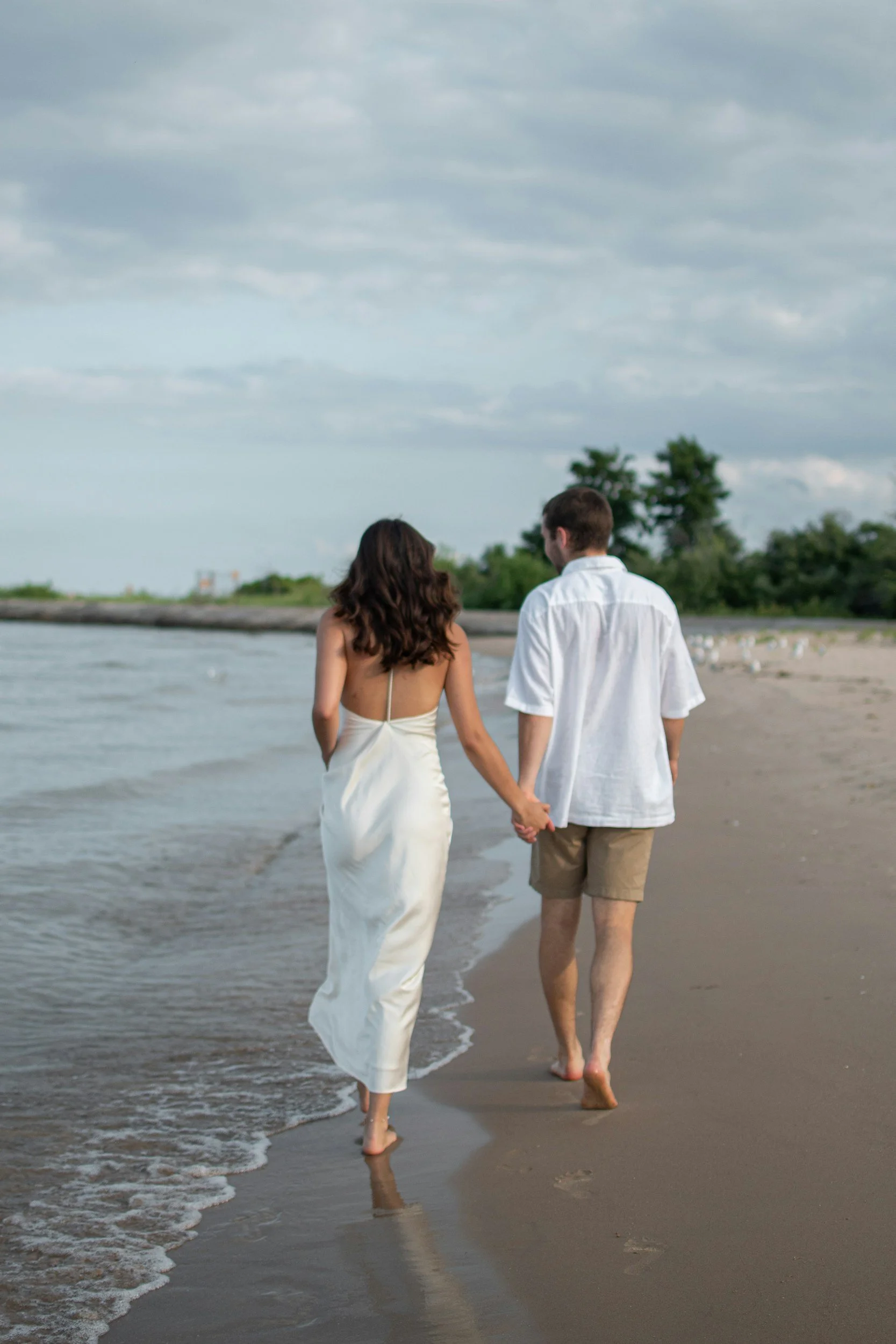 A couple walking hand in hand along the shoreline of a beach, with the woman wearing a white dress and the man in a white shirt and shorts, under a cloudy sky.