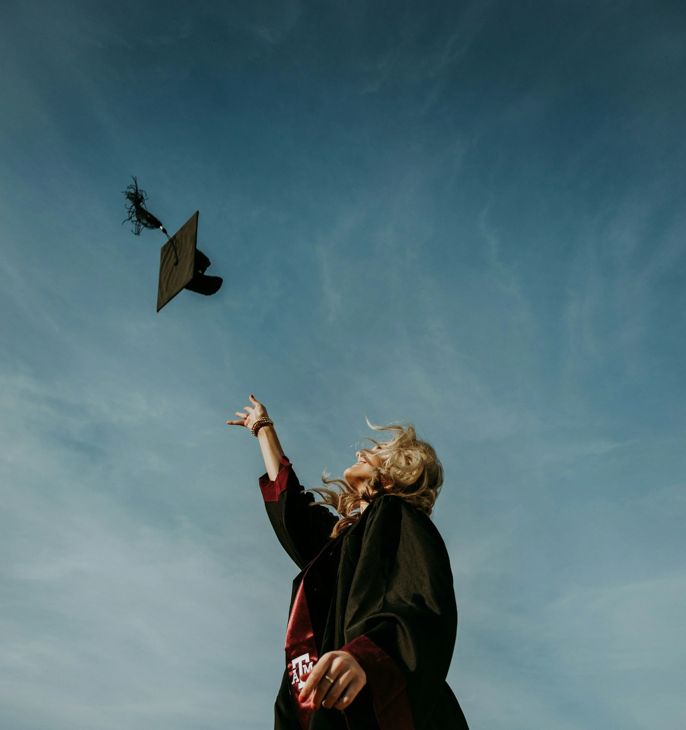 A woman with blonde curly hair raising her arm, wearing a black graduation gown, and holding a small box with an engagement ring inside, against a clear blue sky.