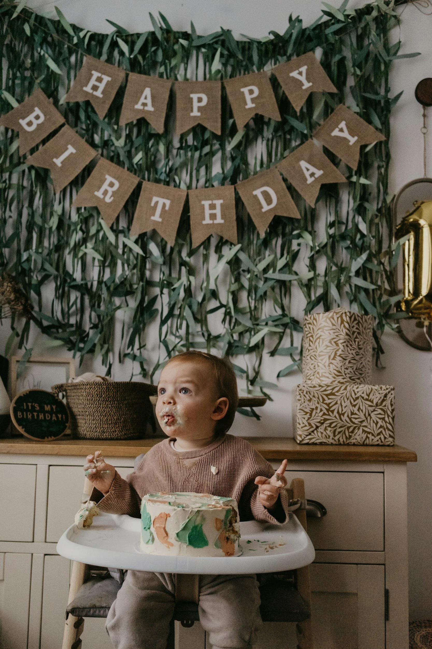 A young child with a piece of birthday cake in front, sitting in a high chair, with cake and frosting on their mouth and hands, celebrating a birthday with a "Happy Birthday" banner in the background.