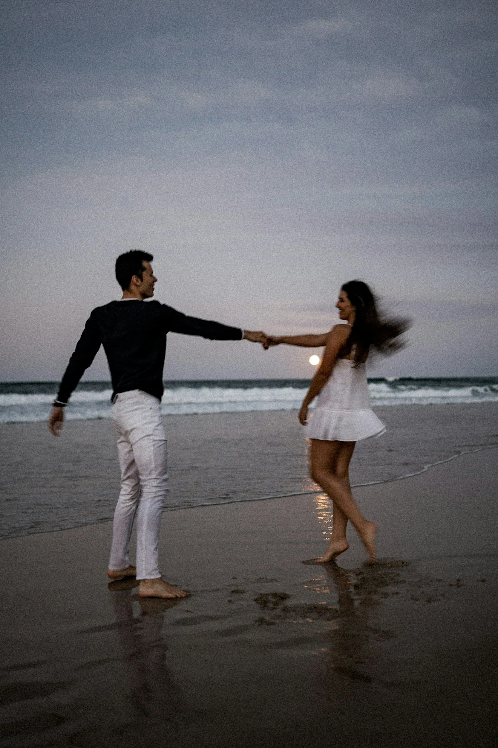 A man and woman holding hands and dancing on the beach at sunset, with waves and a cloudy sky in the background.