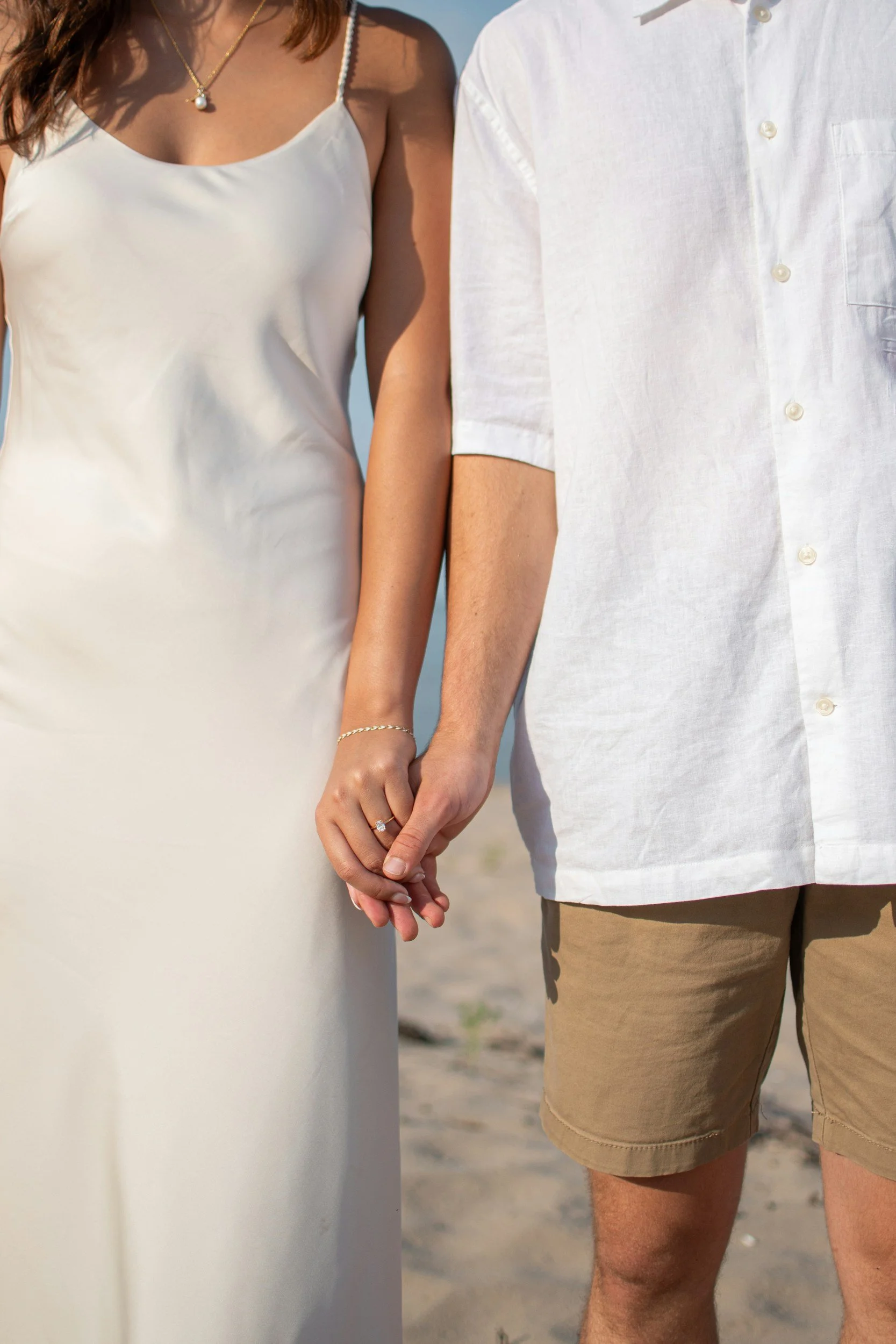 Close-up of a couple holding hands, with the woman wearing a white satin dress and the man in a white short-sleeved shirt and brown shorts, standing on the beach.