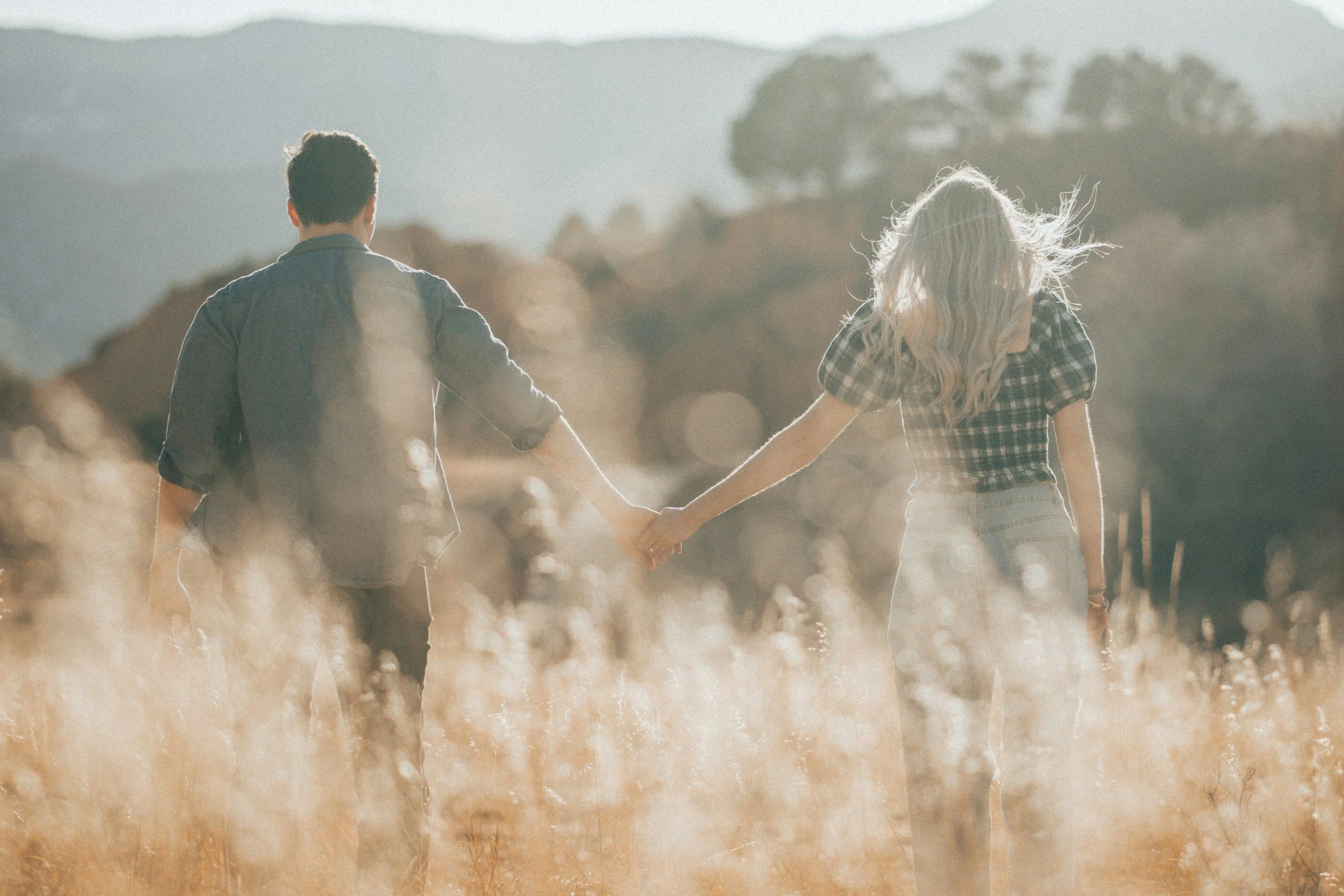 A couple holding hands walking through a field with hills and trees in the background.
