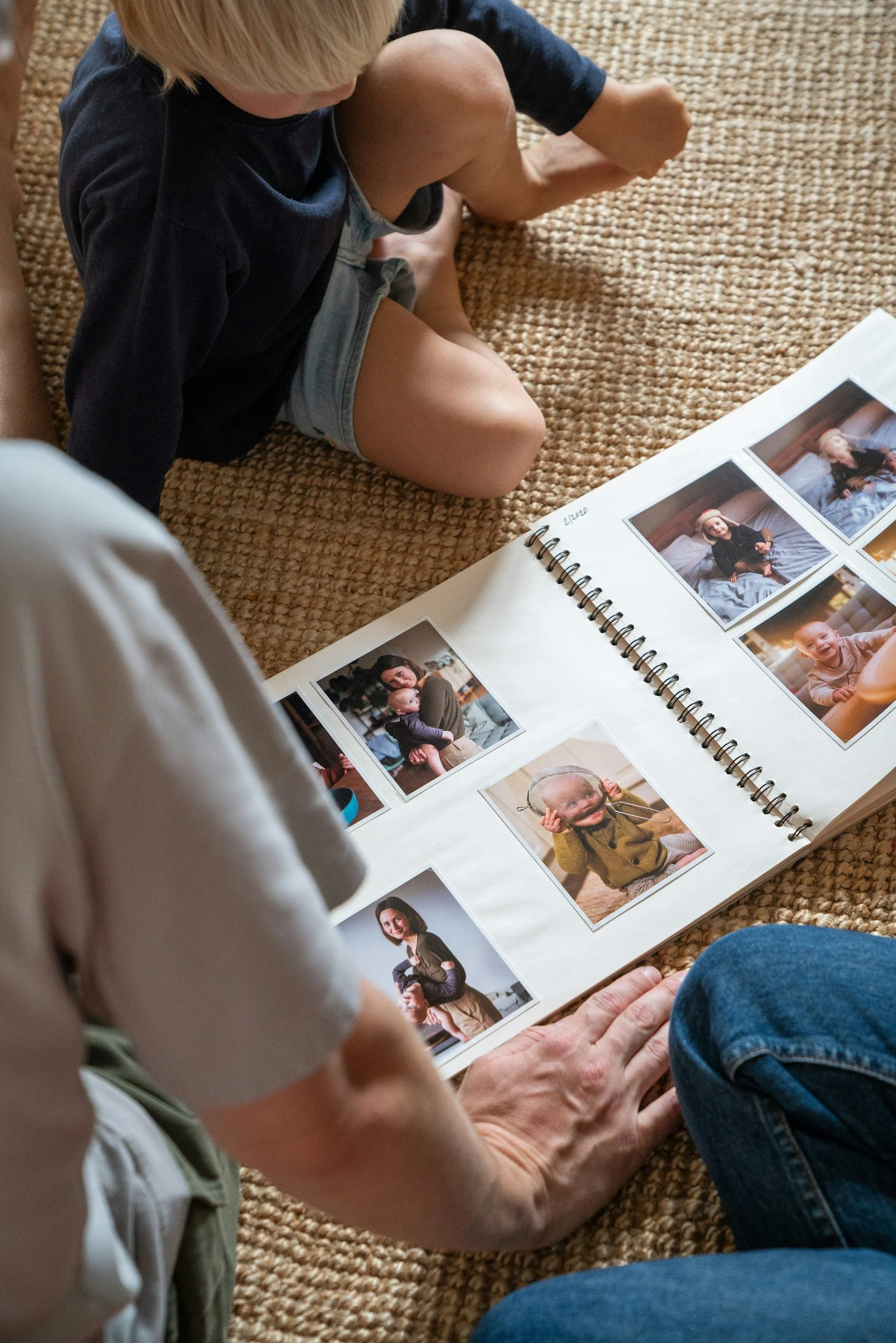 Elderly person and a child looking at a photo album with pictures of children, sitting on a woven rug.