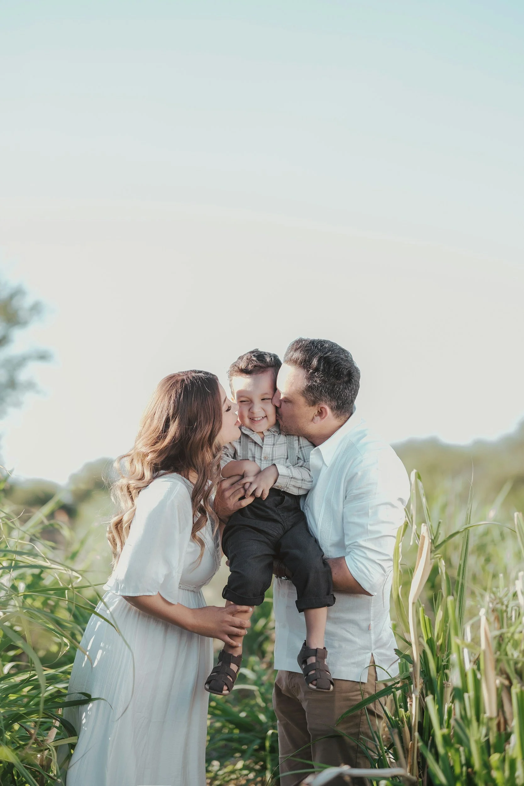 A family of three, mom, dad, and young son, standing in a green outdoor field, sharing a moment of affection and happiness.