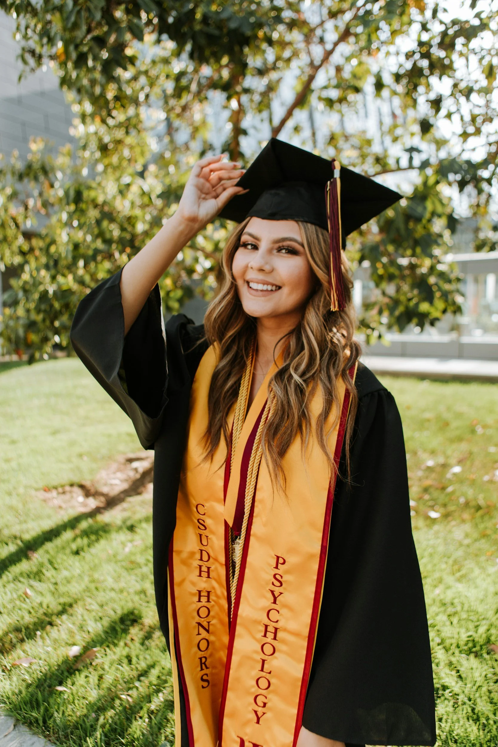 A young woman in a black graduation gown and cap, wearing a gold honor sash, outdoors underneath a tree, smiling at the camera.
