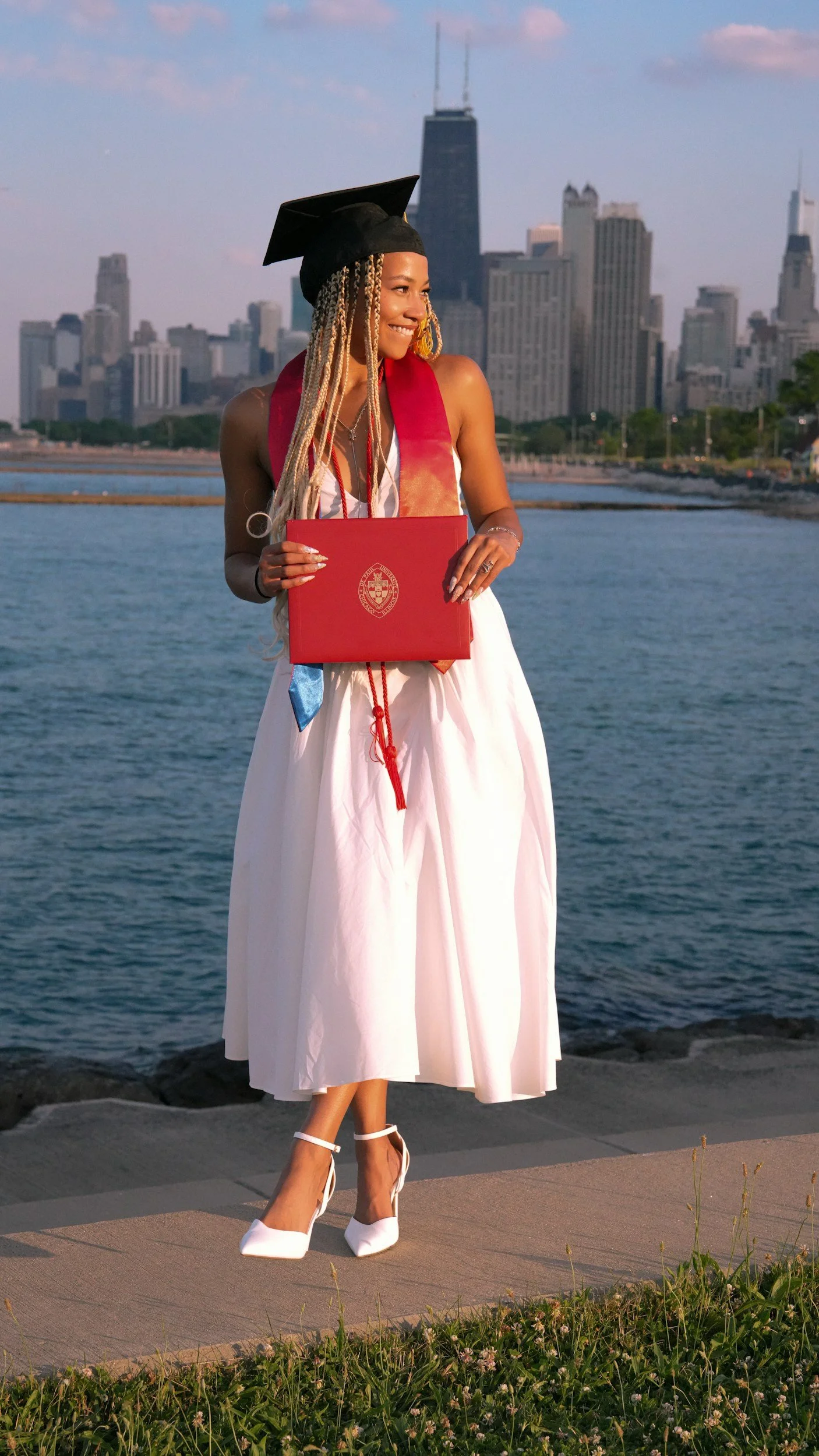 A woman in a white dress and white heels with ankle straps, wearing a graduation cap and red stole, holding a diploma or certificate, standing outdoors near water with a city skyline in the background.