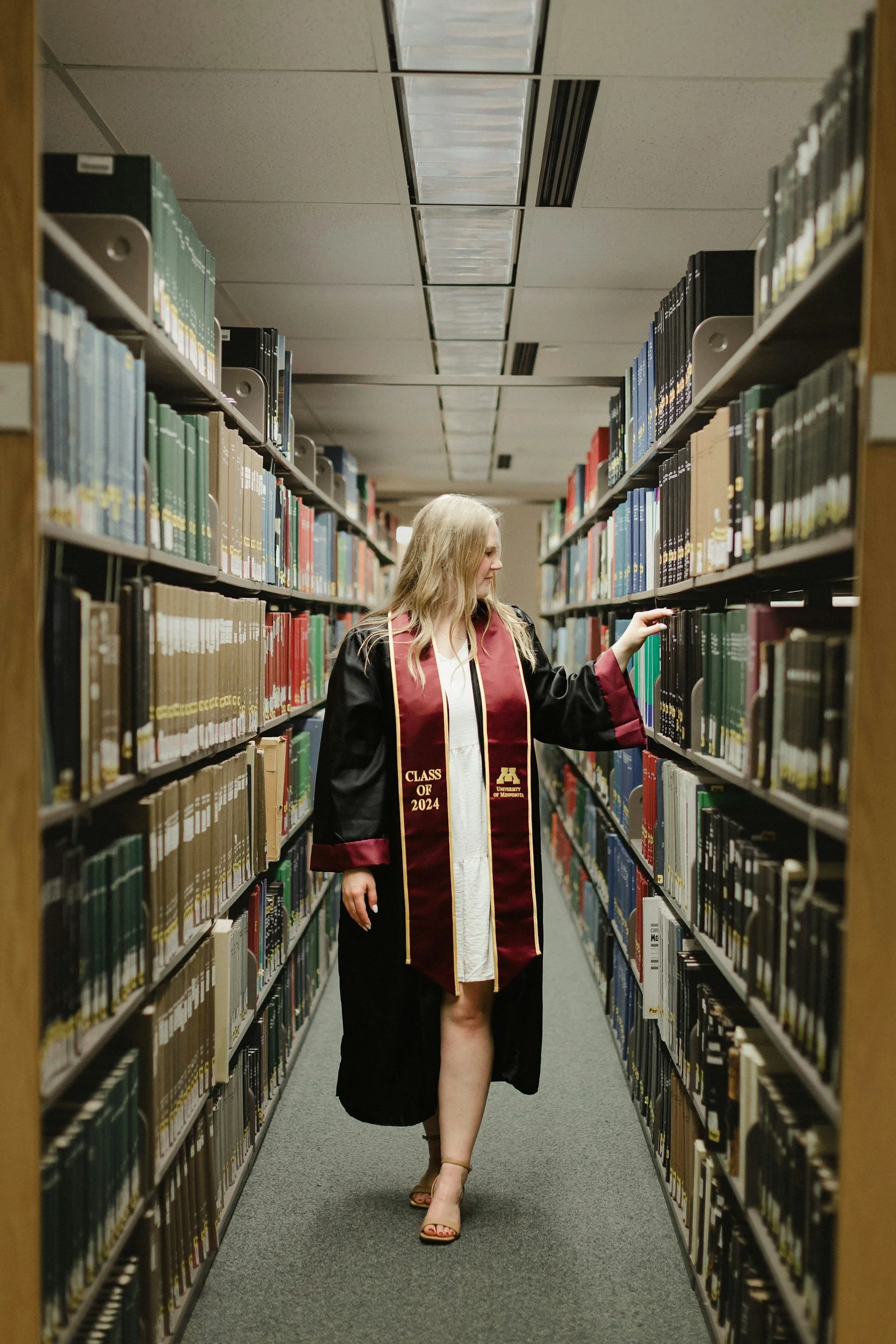 A woman wearing a graduation gown with 'Class of 2024' and the University of Minnesota logo, walking through a library aisle lined with bookshelves.