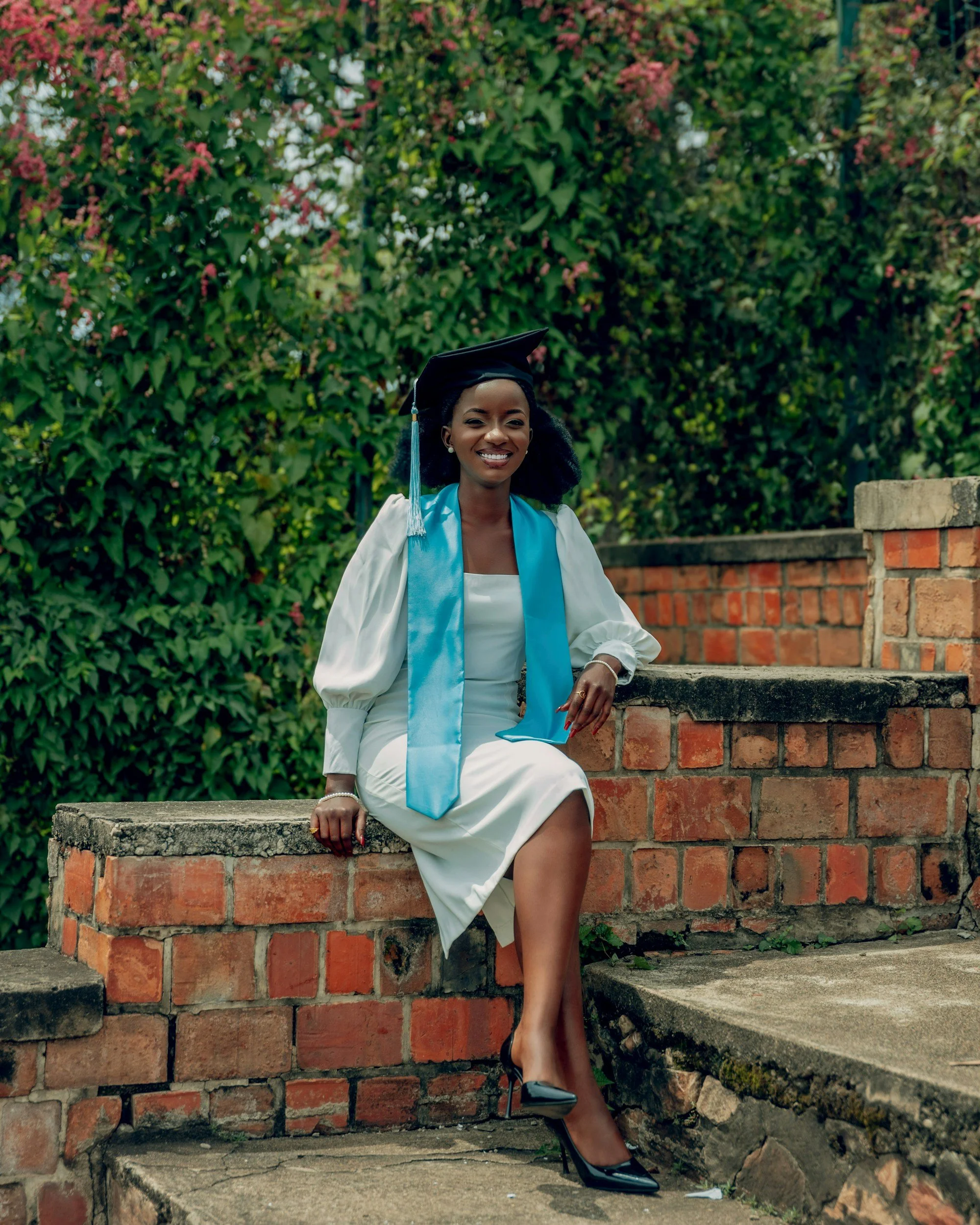 A young woman in a white dress and blue sash is sitting on a brick ledge in front of a green leafy background. She is wearing a graduation cap and smiling at the camera.