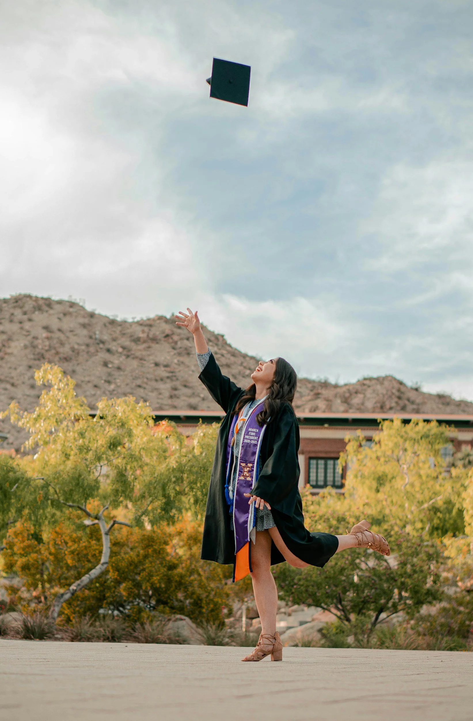 A young woman in a graduation gown and cap performing a yoga pose outdoors. She is standing on one leg, with the other leg bent back and held by her hand, while tossing her cap in the air. The background features trees with autumn foliage, a building, and a mountain with a cloudy sky.