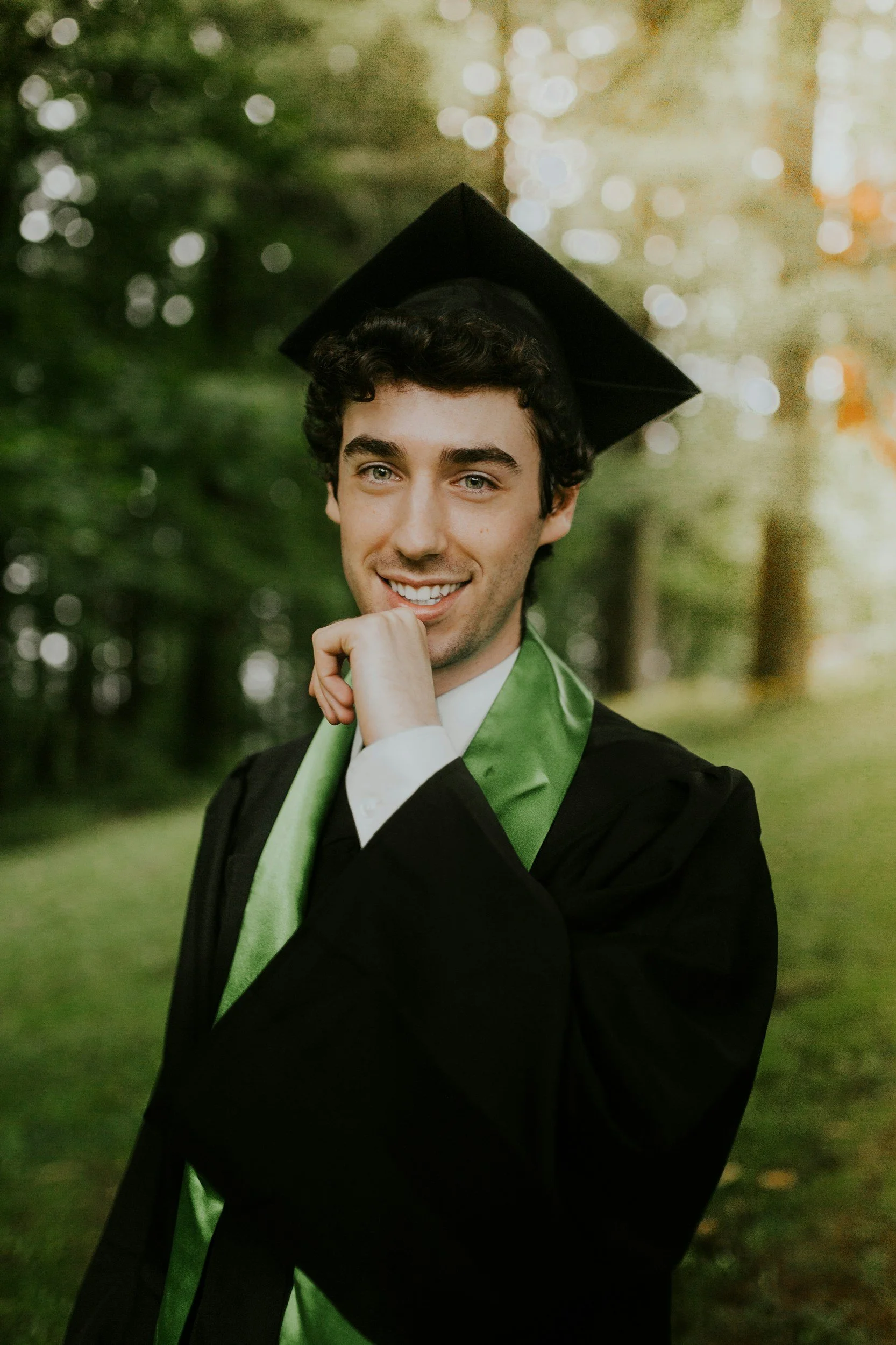 Young man in a graduation cap and gown with a green sash, standing outdoors with trees and sunlight in the background.