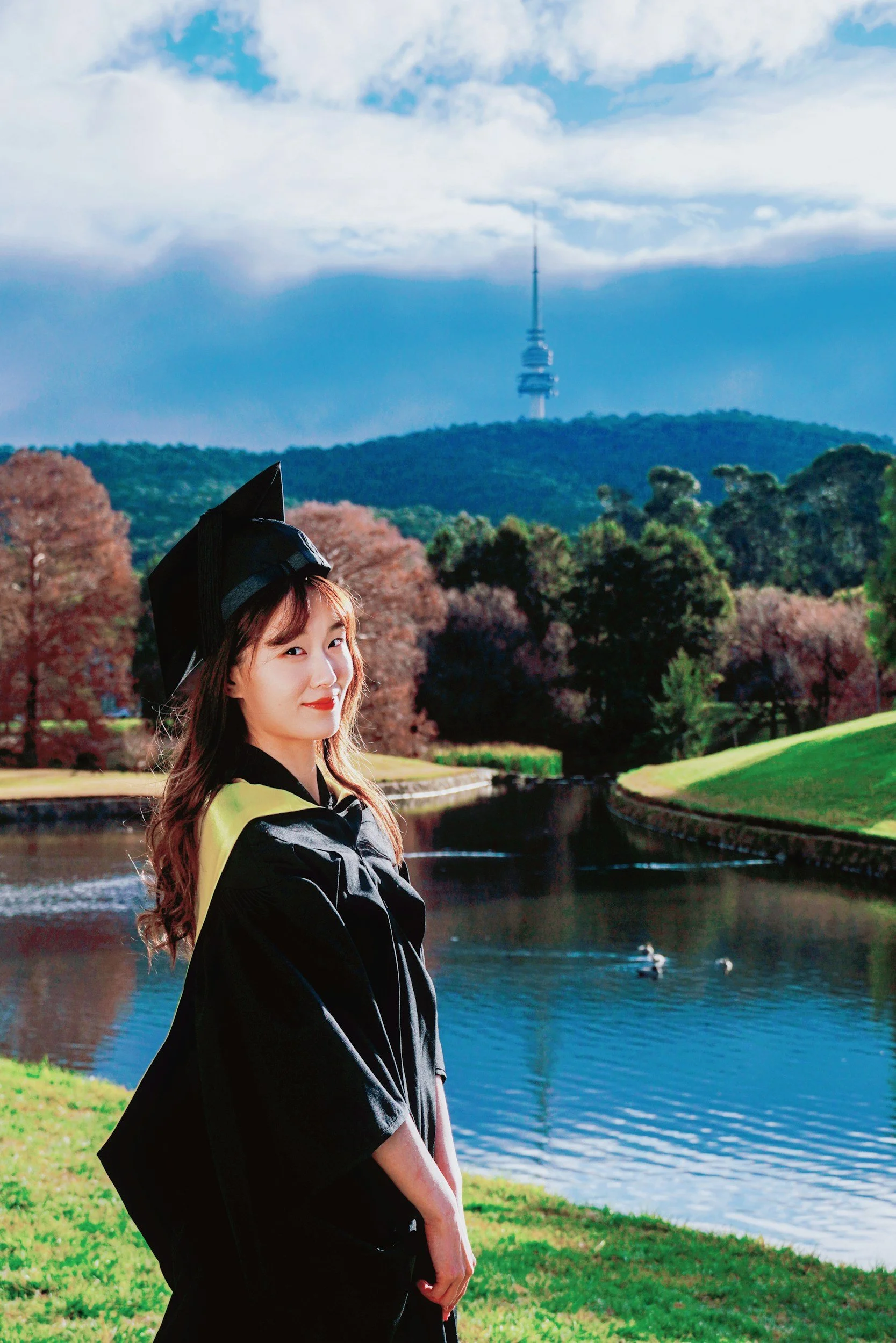 A young woman dressed in her graduation cap and gown standing outdoors with a river, ducks, green trees, and a large tower in the background.