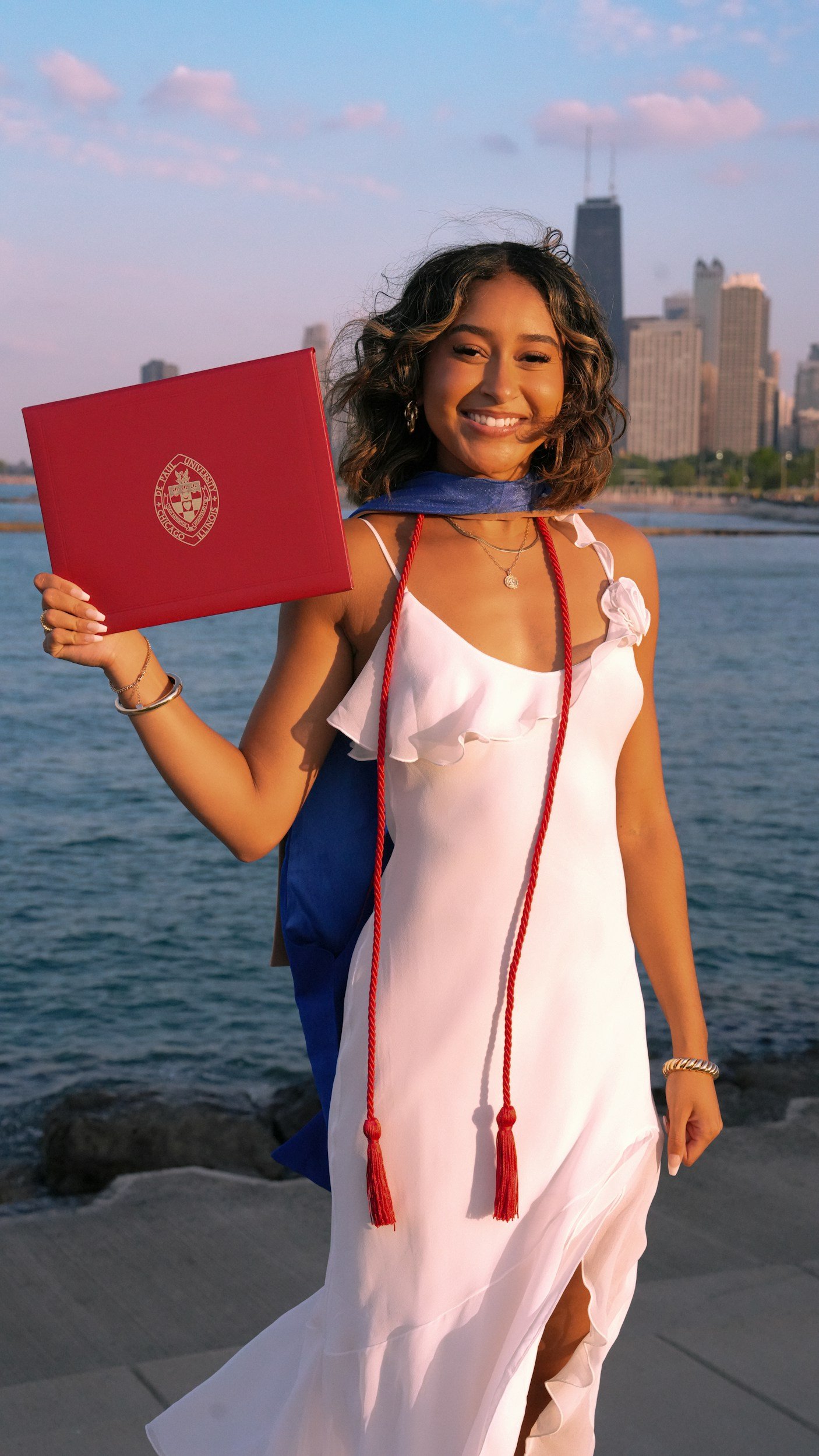 Young woman in a white dress holding a red diploma, smiling outdoors near water with a city skyline in the background.
