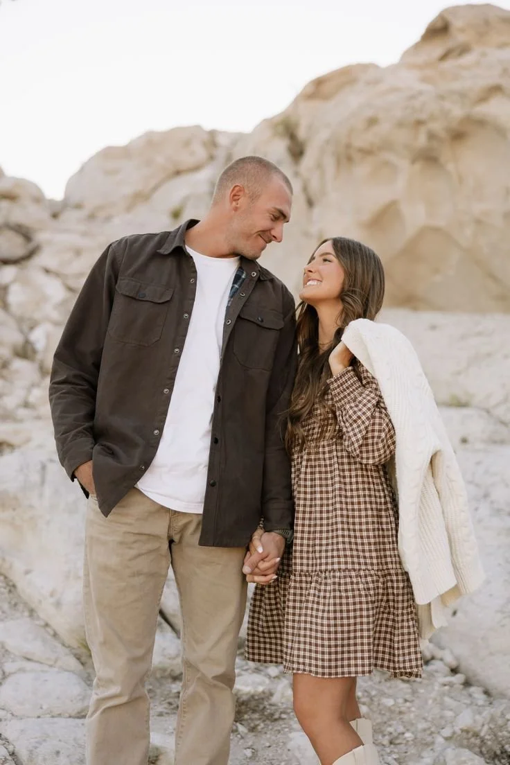 A man and woman holding hands, smiling at each other in a rocky outdoor setting with beige rock formations.