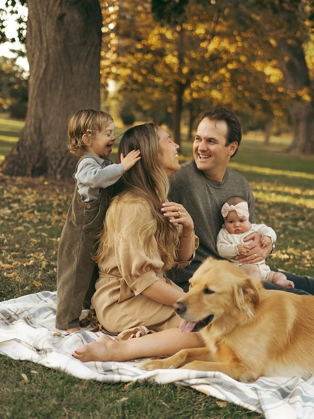 Family of four with a dog sitting on a blanket outdoors in a park, surrounded by trees with autumn leaves, smiling and interacting.